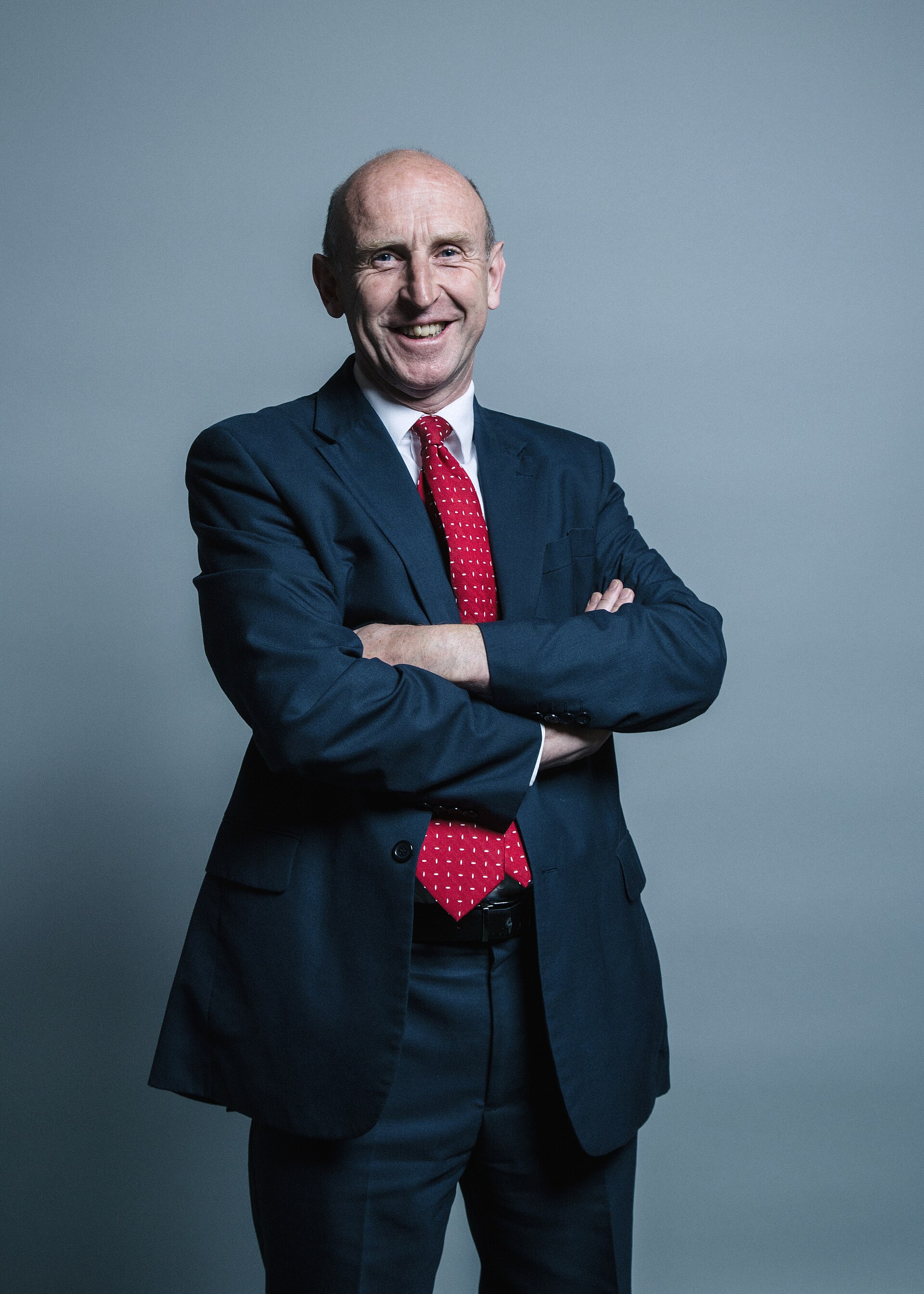 A middle-aged man in a dark blue suit, red tie, and white shirt, with his arms crossed, standing against a gray background.