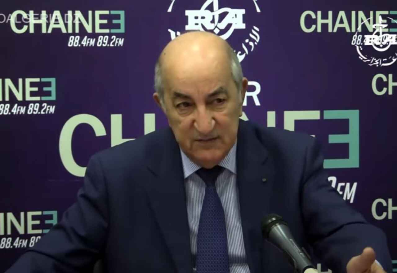 A balding older man in a dark blue suit, light blue shirt, and dark blue tie speaks into a microphone in front of a purple backdrop with logos and text.