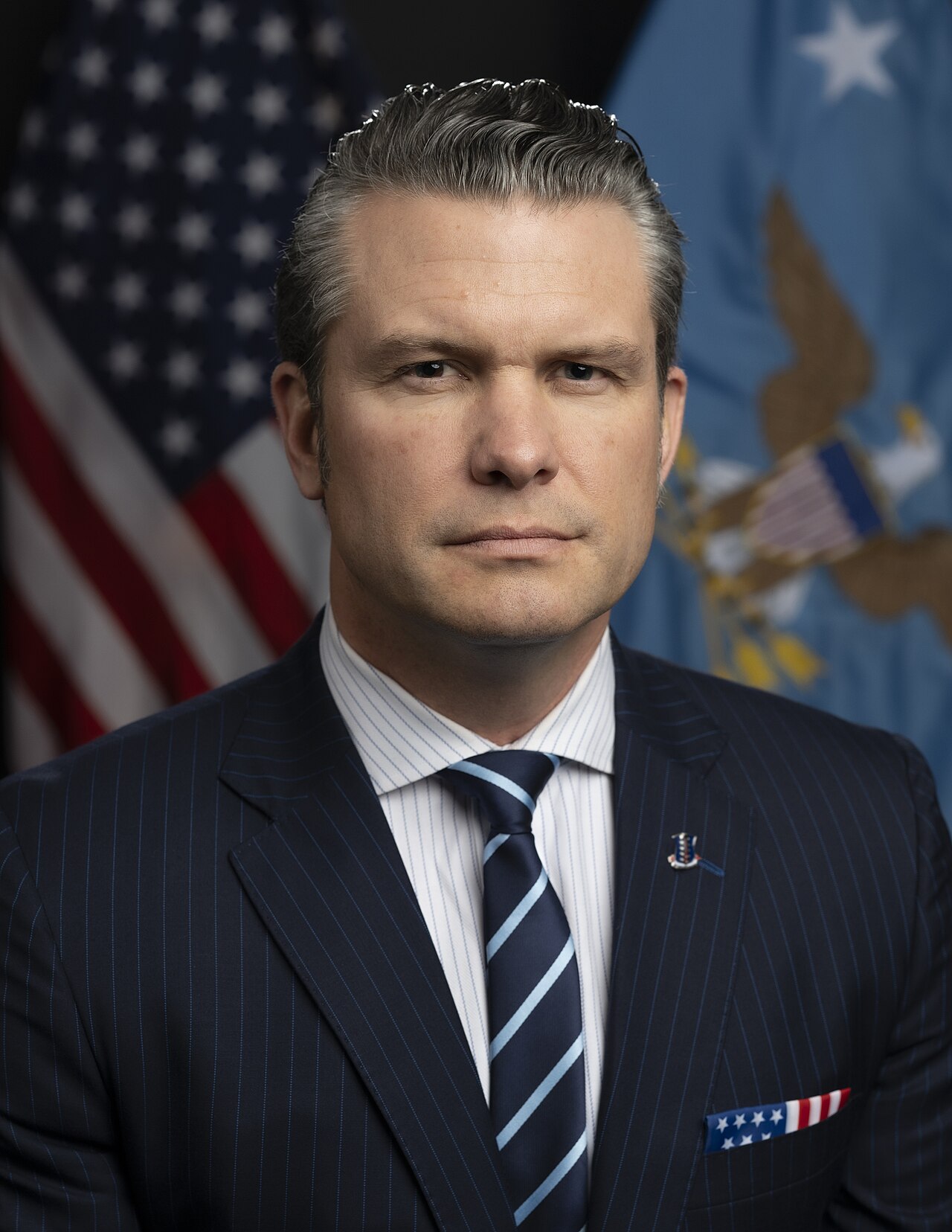 Pete Hegseth, a middle-aged man with short graying hair, wearing a navy blue pinstripe suit, white shirt, and navy blue tie with light blue stripes, with an American flag pin on his lapel and a matching pocket square, standing in front of two flags.