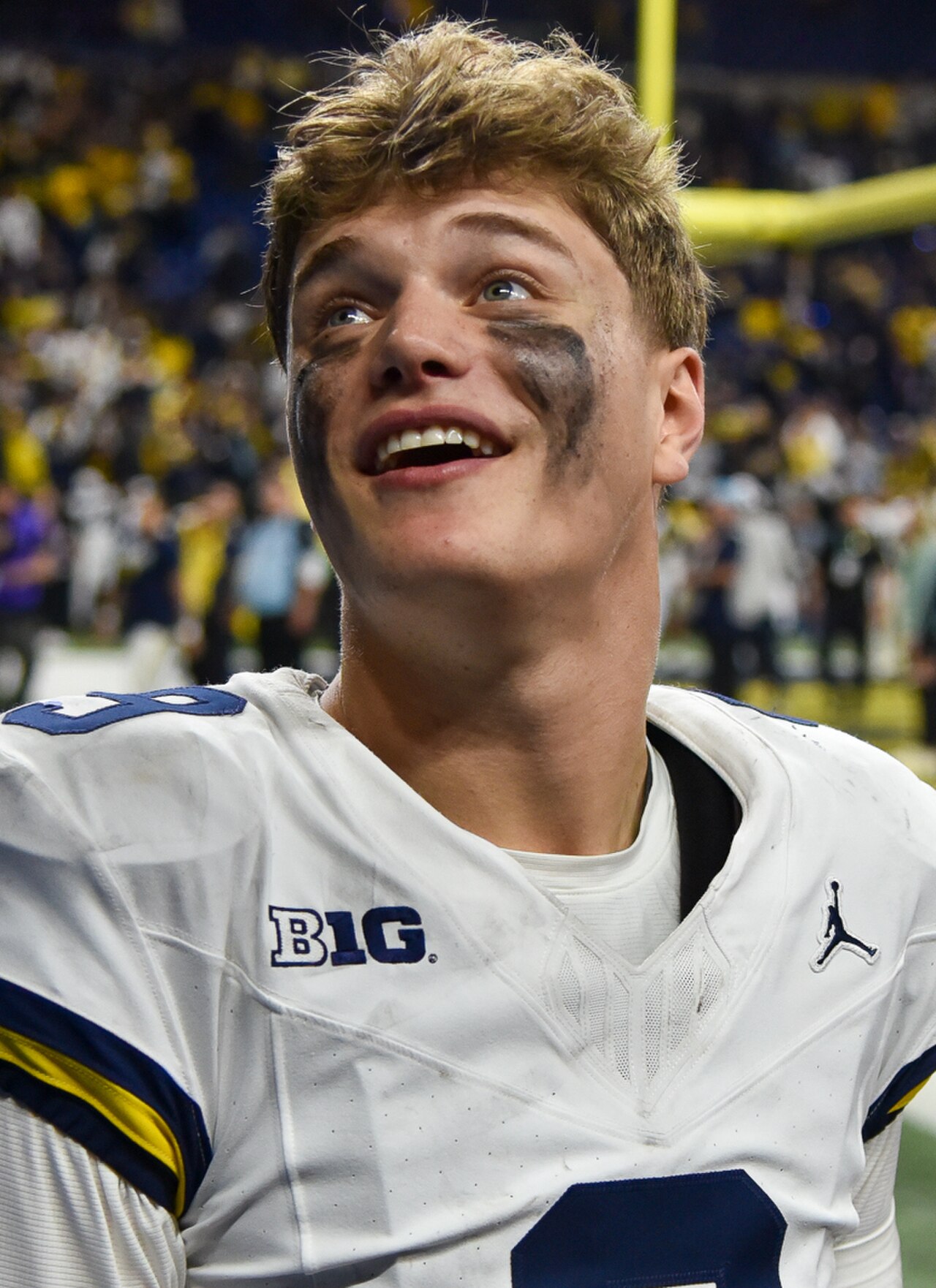 A smiling young man in a white football jersey with blue and yellow stripes, a Jordan logo, and BIG text on the chest, with black face paint.