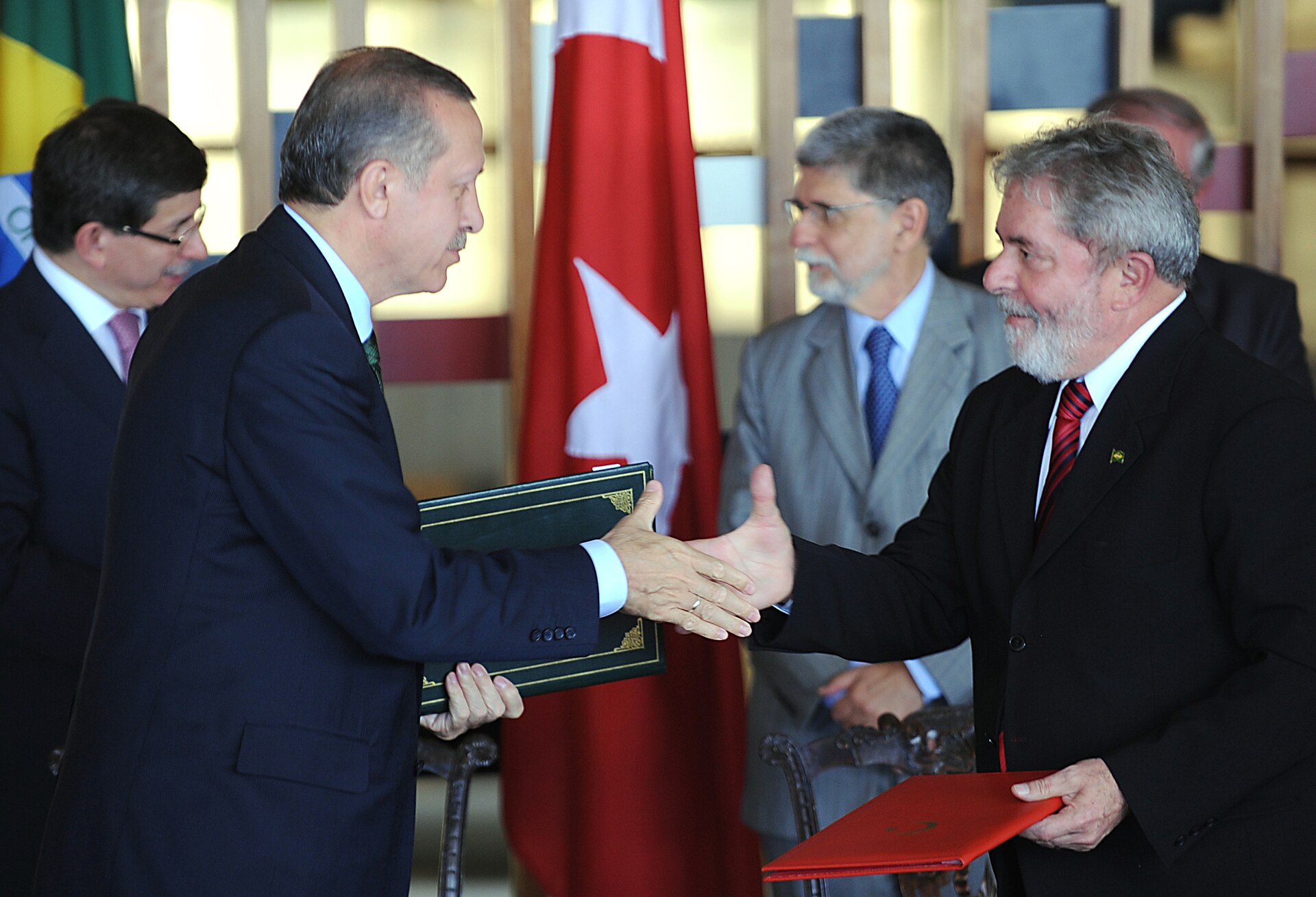 Two men in suits shaking hands, one with a red folder and the other with a green binder, with other men and flags in the background.
