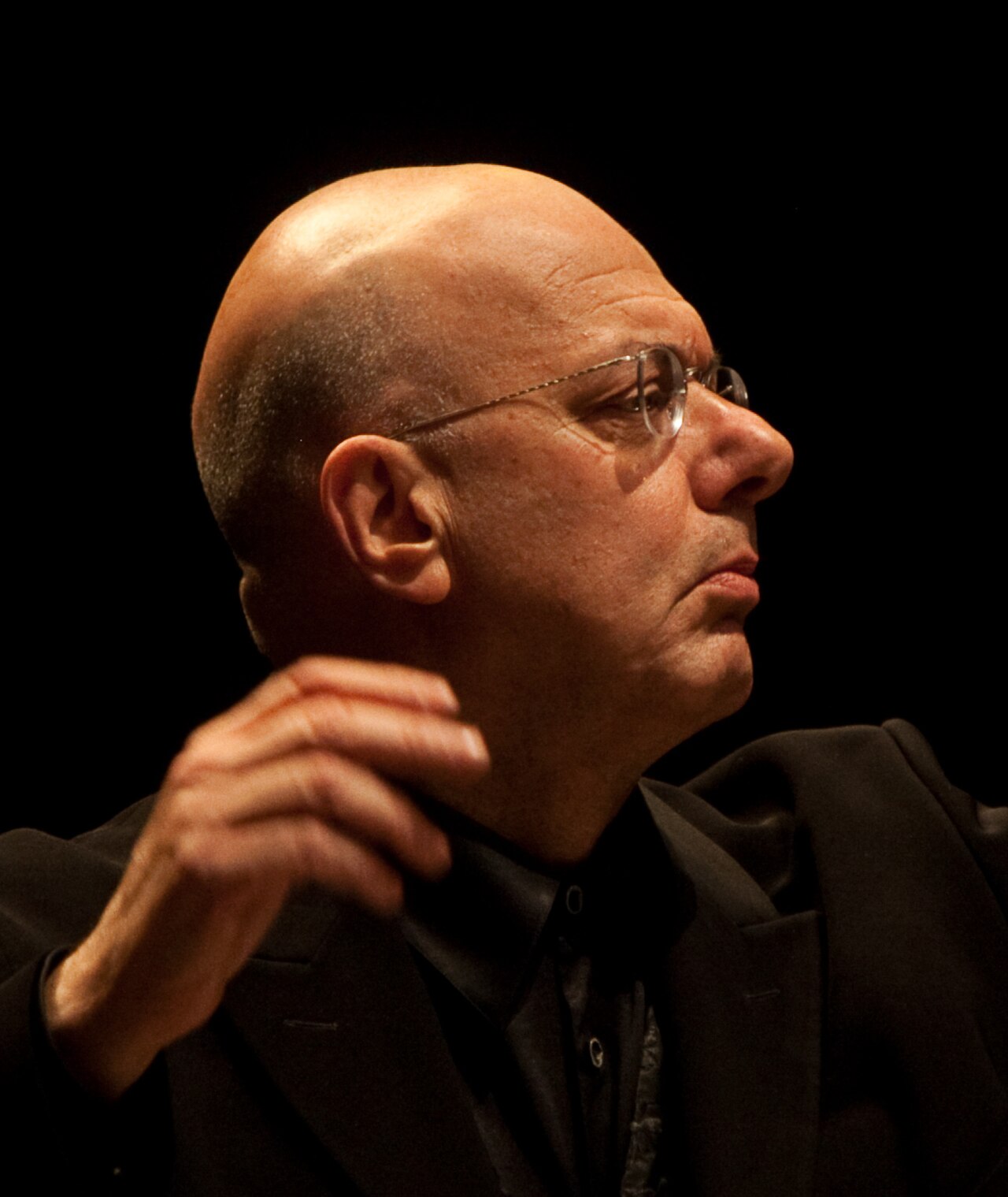 Leon Botstein, a bald man with glasses and a black suit, looking to the right with his hand raised near his shoulder.