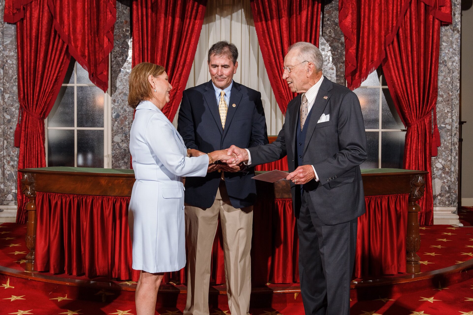 Three people in formal attire stand in front of red curtains and a wooden table.