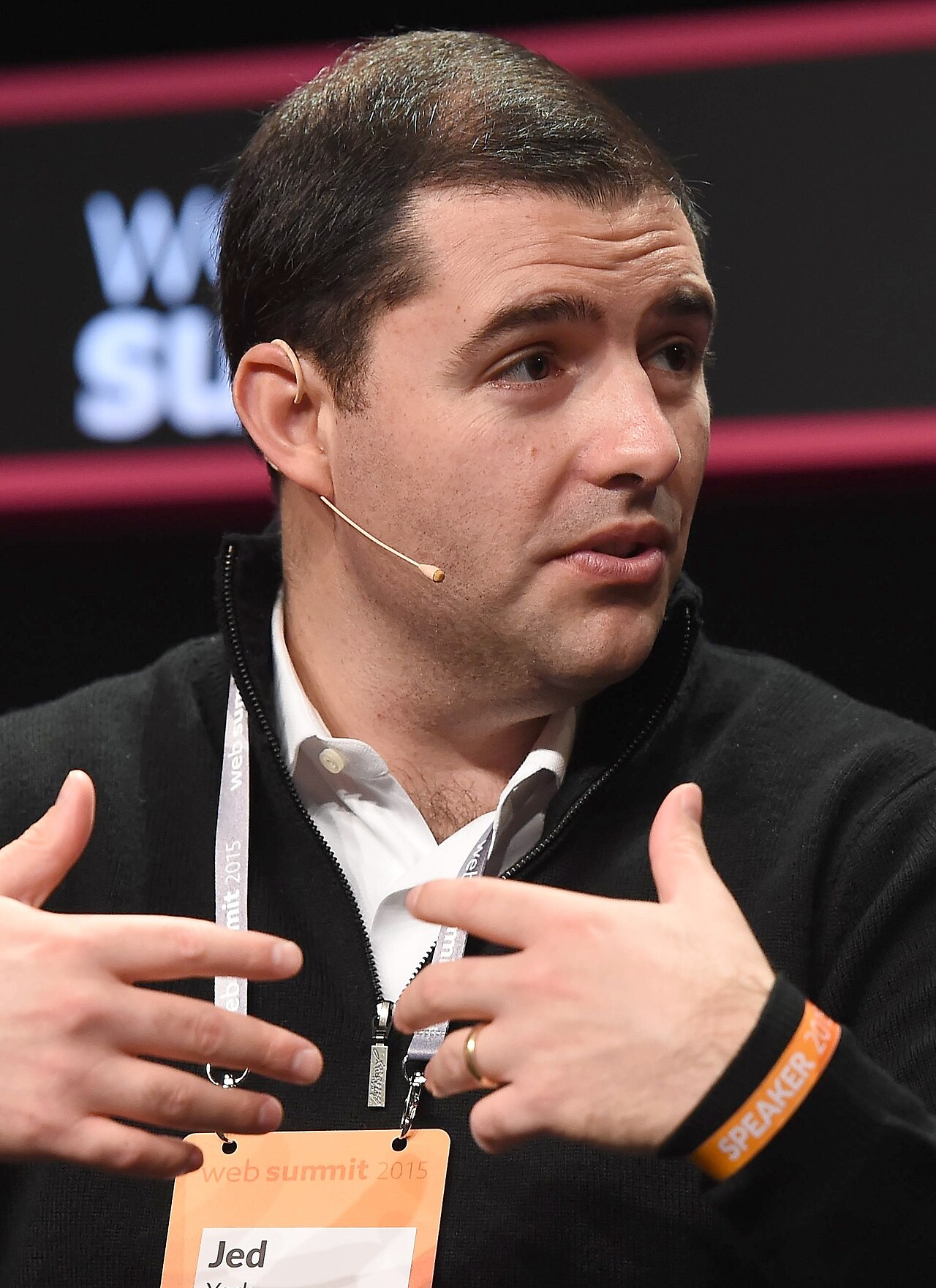 A man with short dark hair, wearing a black sweater over a white collared shirt, a lanyard with an orange badge that reads 'Jed', and an orange wristband that reads 'SPEAKER', gesturing with his hands.