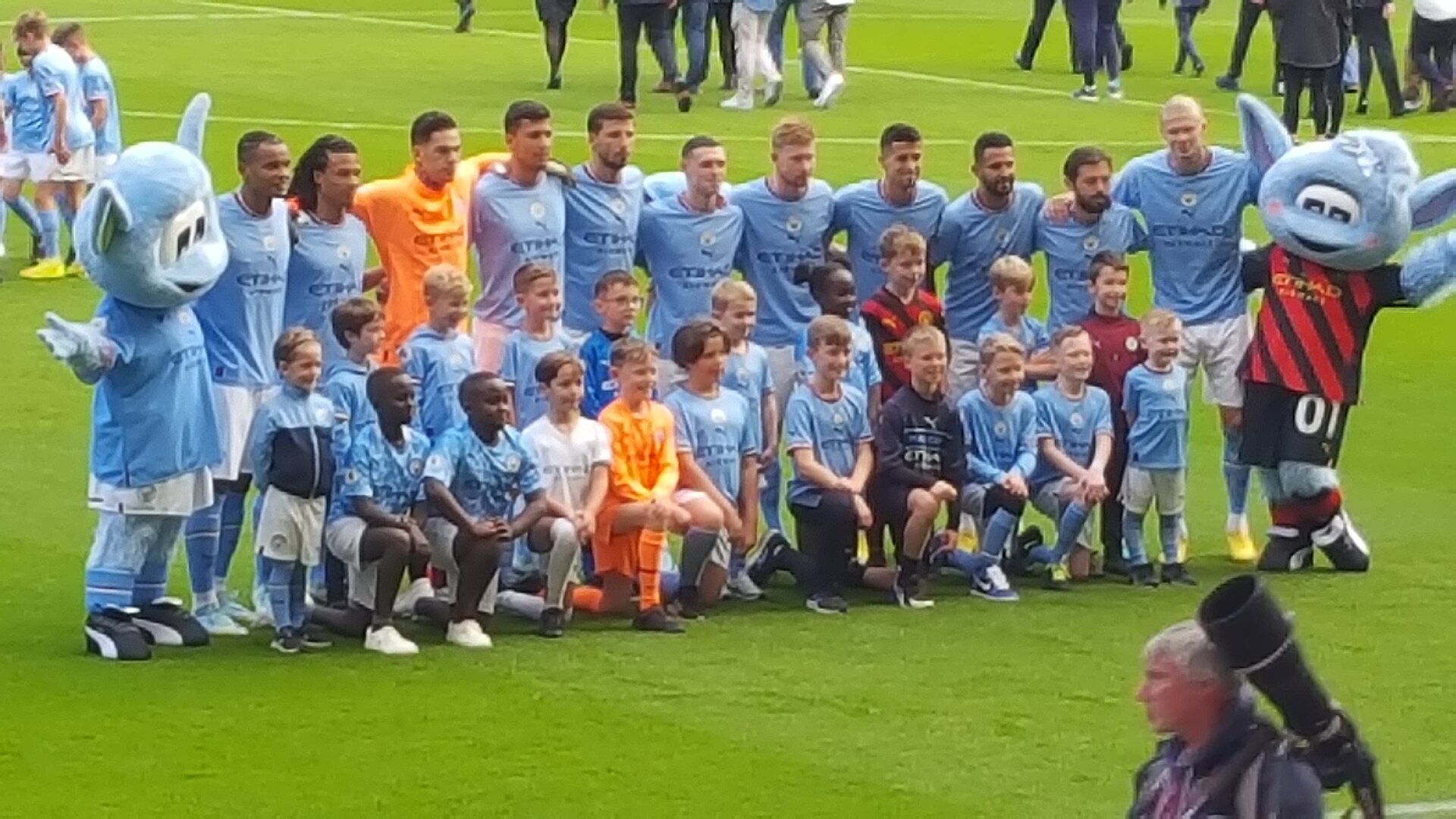 A group of Manchester City players and mascots pose for a photo on a green field.