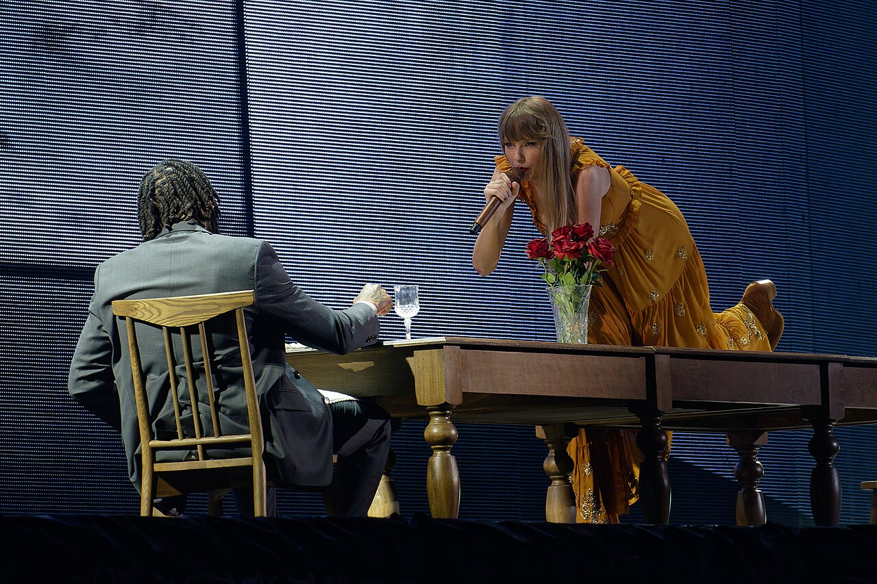 Taylor Swift sings into a microphone in a gold gown beside a pianist onstage.