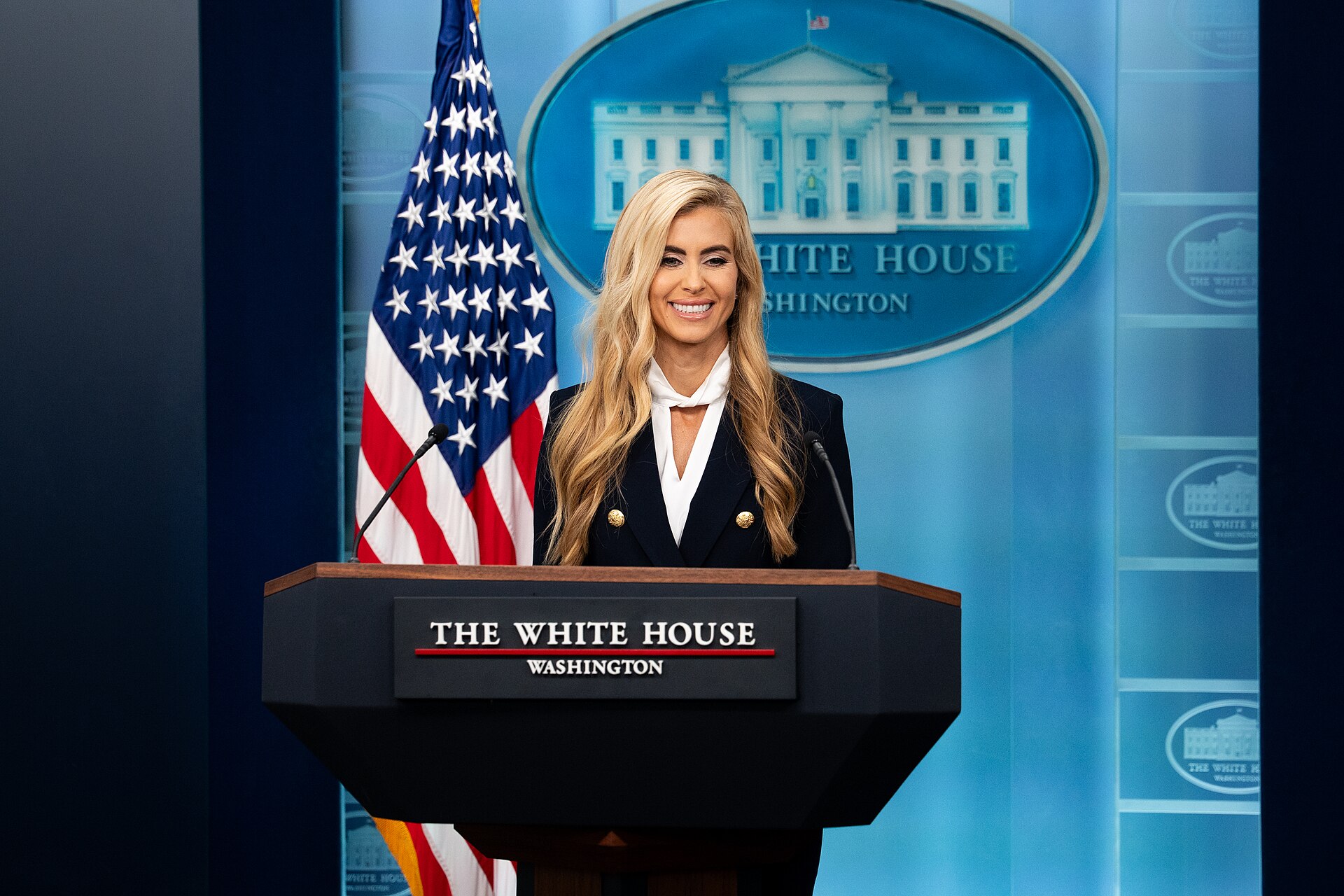 A woman with long blonde hair, wearing a dark blazer over a white shirt and scarf, smiles while standing behind a podium with "THE WHITE HOUSE WASHINGTON" printed on it. An American flag is visible to her left.