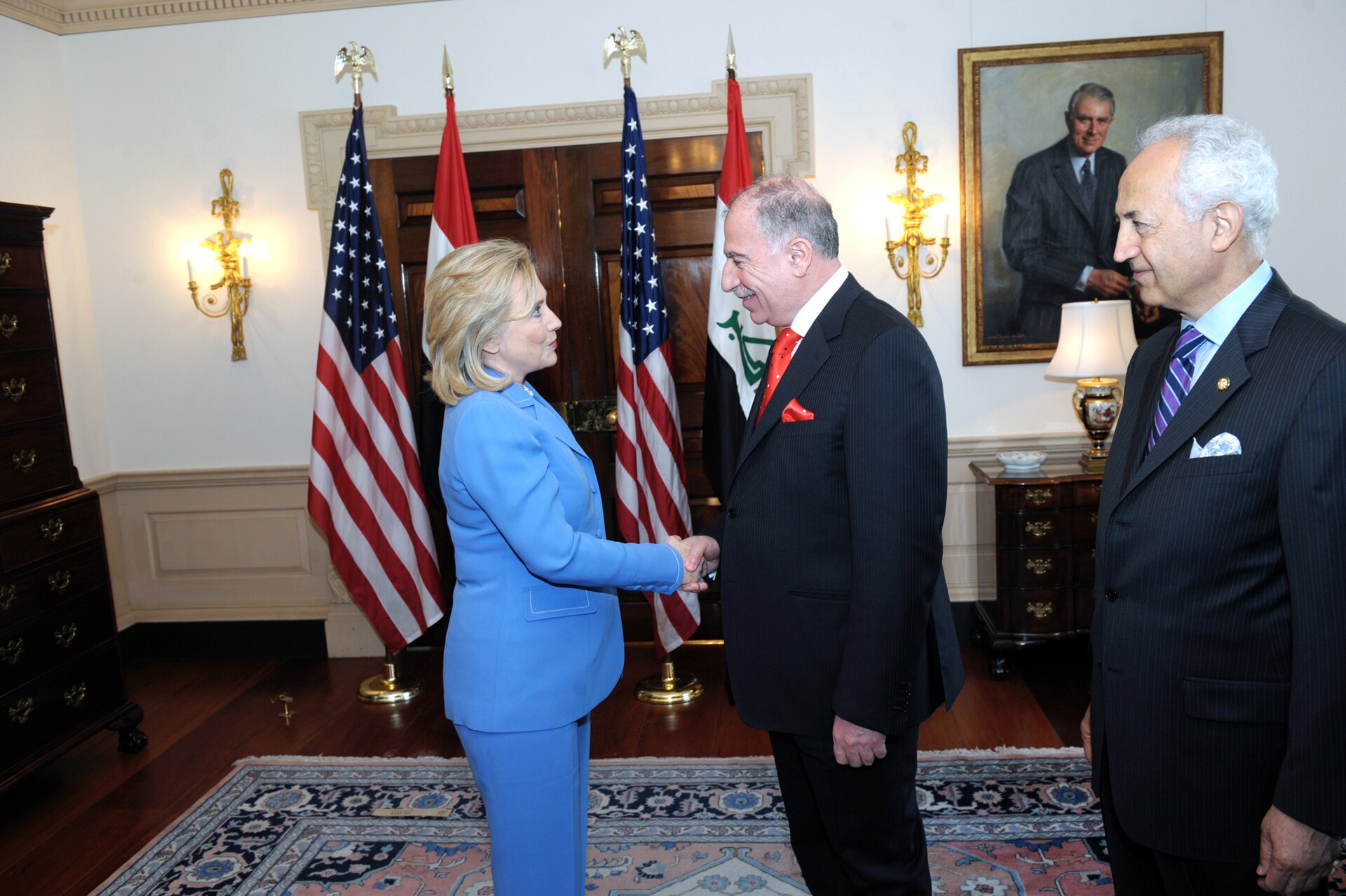 A woman in a blue suit shakes hands with a man in a dark suit with a red tie, while another man in a pinstripe suit stands to the right. Two American flags and an Iraqi flag are visible behind them, along with a portrait of a man in a suit on the wall.