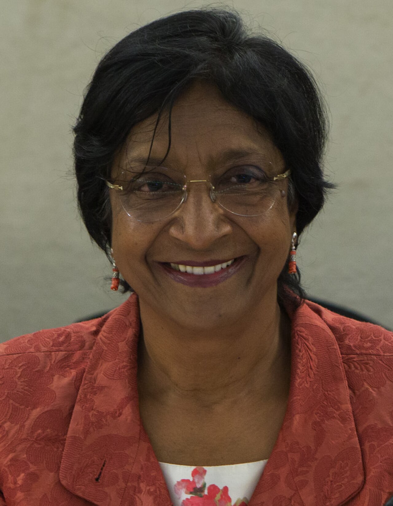 A woman with short black hair, wearing glasses, a red jacket, and a white top with pink flowers, smiling at the camera.