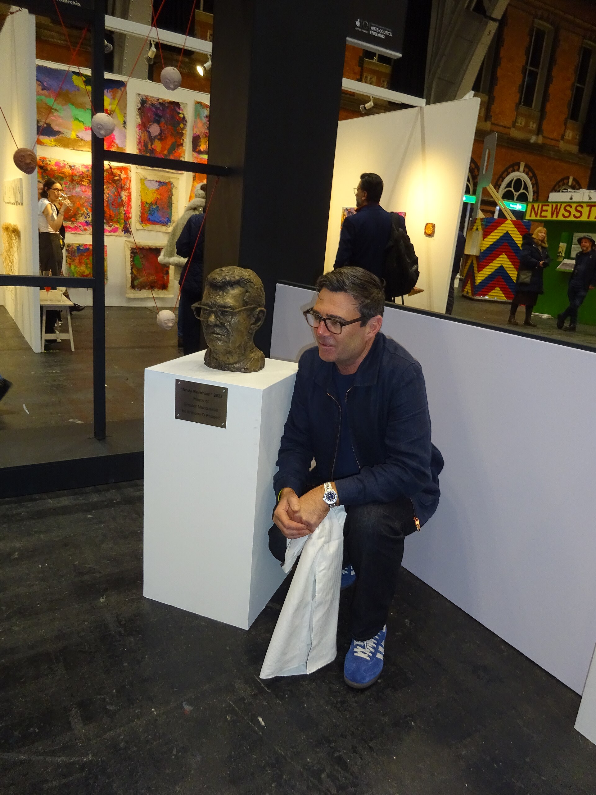 A man in glasses and a dark jacket kneels next to a bronze bust of a man wearing glasses, with a plaque on the pedestal reading "Andy Burnham 2025 Mayor of Greater Manchester".
