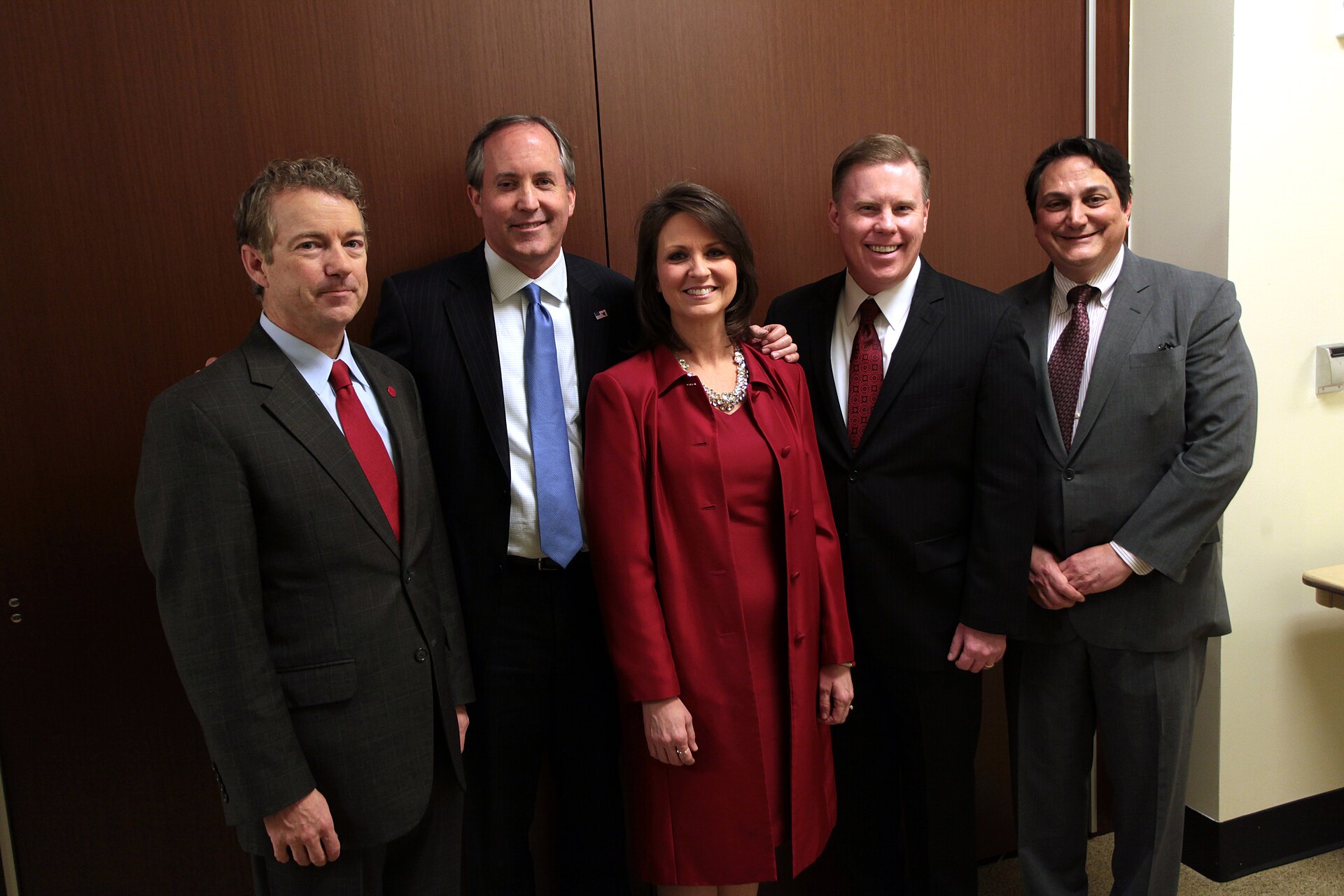 Five people, four men and one woman, stand in a line against a wood-paneled wall. The man on the far left wears a dark suit and a red tie. The next man wears a dark suit and a blue tie. The woman in the center wears a red coat and dress with a statement necklace. The man to her right wears a dark su