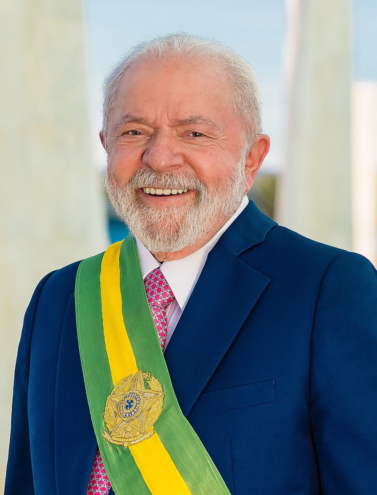 Luiz Inácio Lula da Silva, wearing a blue suit, white shirt, pink tie, and a sash with the colors of Brazil, smiles at the camera.