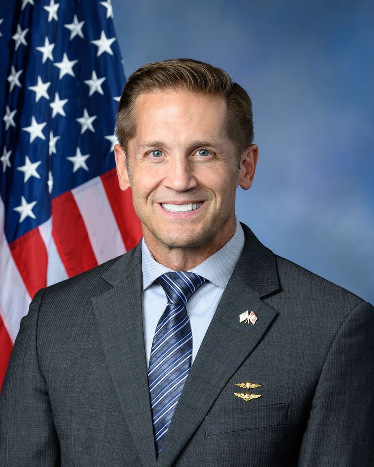 A smiling man in a suit with a US flag behind him