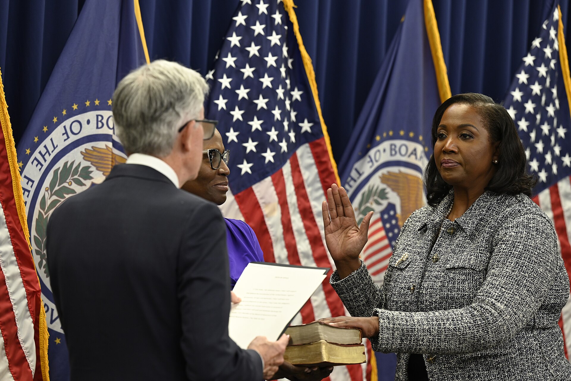 A woman in a tweed jacket places her right hand on a stack of books while raising her left hand, as if taking an oath. She is standing in front of American flags and flags with the seal of the Board of Governors of the Federal Reserve System.