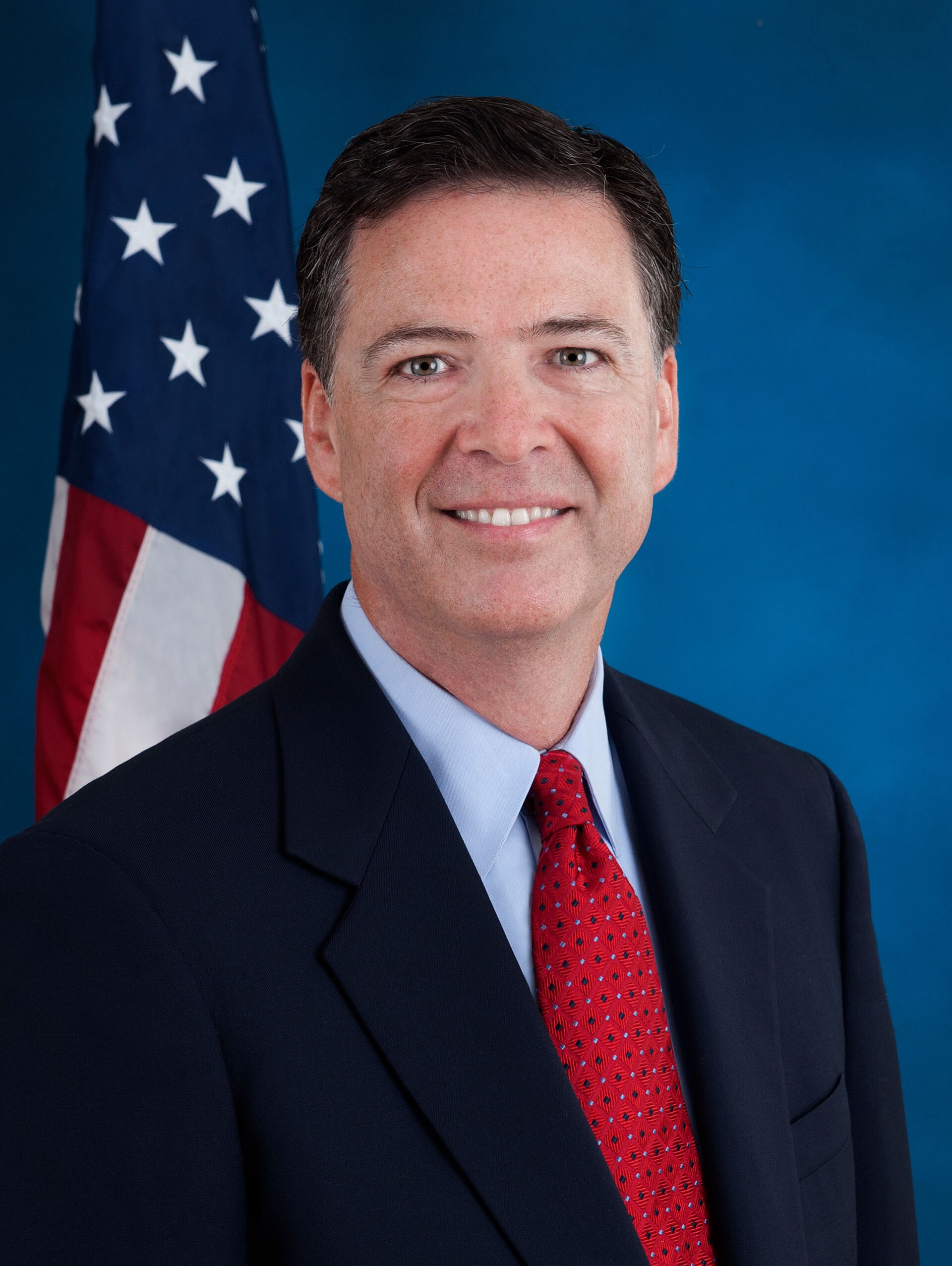 James Comey smiles in a suit and red tie, with an American flag behind him.