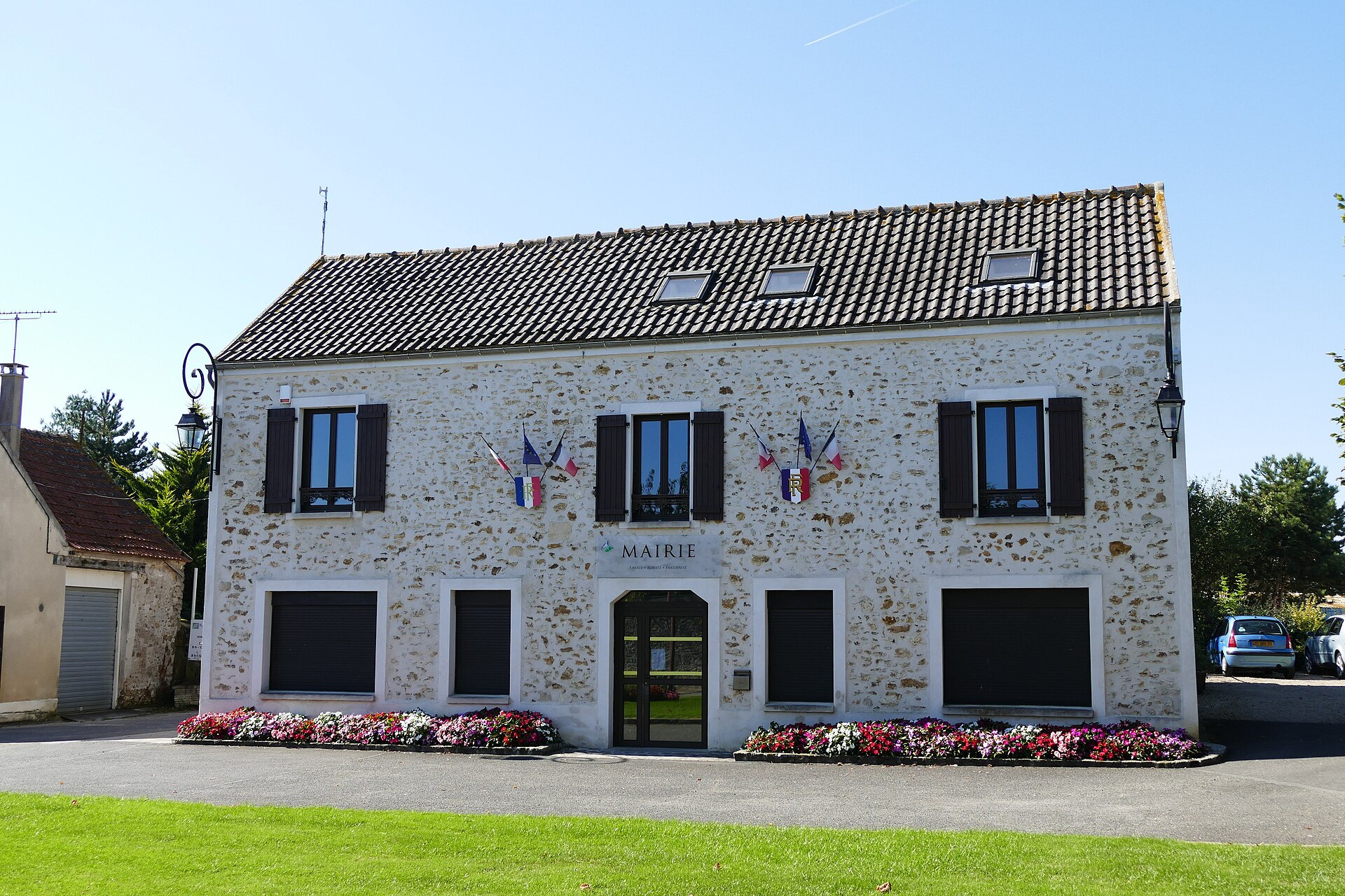 A stone building with a tiled roof, labeled "MAIRIE" above the entrance, with French flags displayed on either side of the central window. Flower beds line the front of the building, and a grassy lawn is in the foreground.