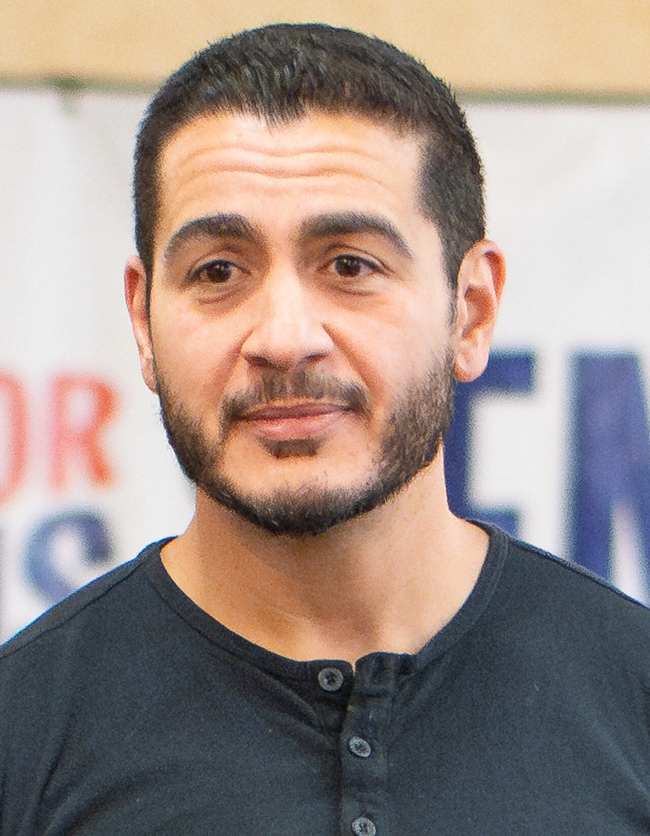 A close-up headshot of a man with dark hair, a beard, and a mustache, wearing a black henley shirt.