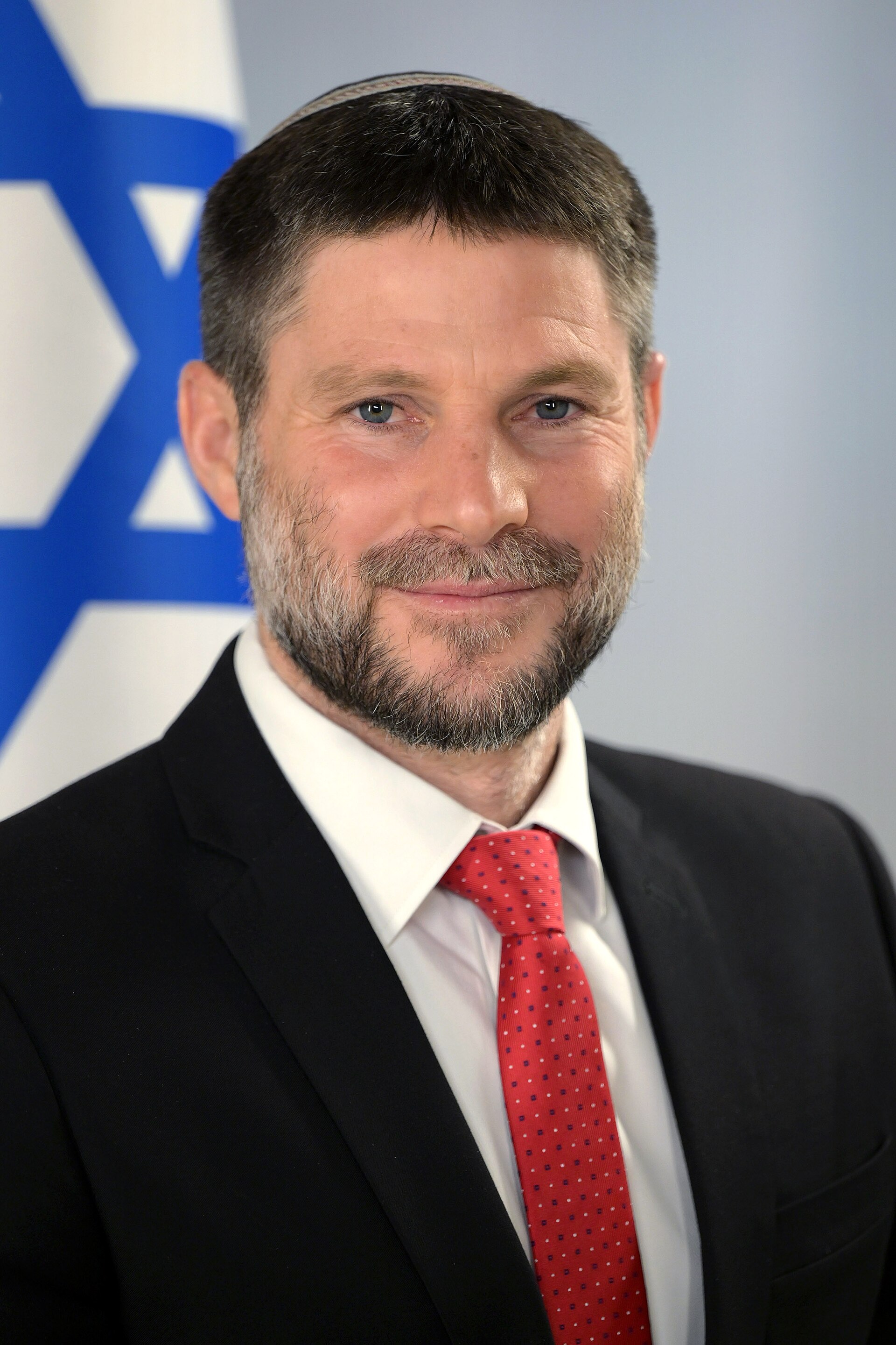 A man with short dark hair, a beard, and a yarmulke, wearing a black suit, white shirt, and red tie, stands in front of a flag featuring a blue Star of David on a white background.