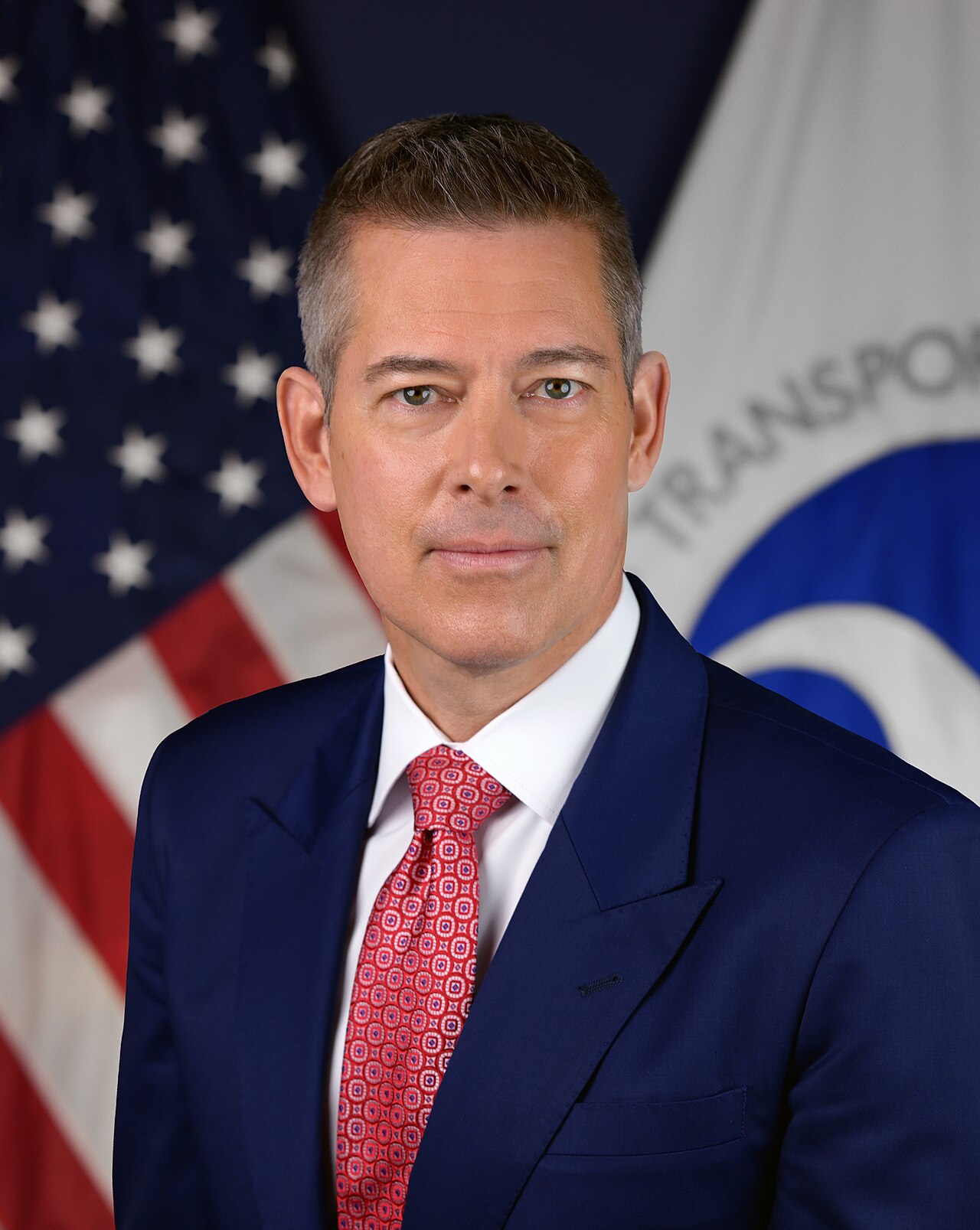 Sean Duffy in a navy suit and red tie standing in front of an American flag and a Department of Transportation flag.