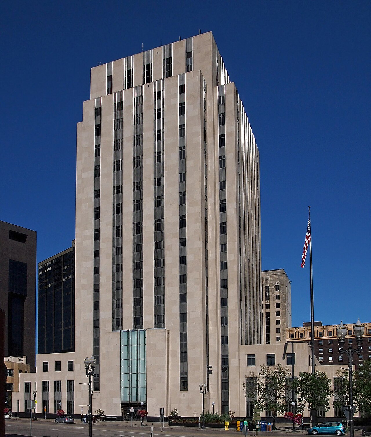 A tall, light-colored stone building with many windows stands against a clear blue sky. An American flag flies from a flagpole to the right of the building. Other buildings and trees are visible in the background.