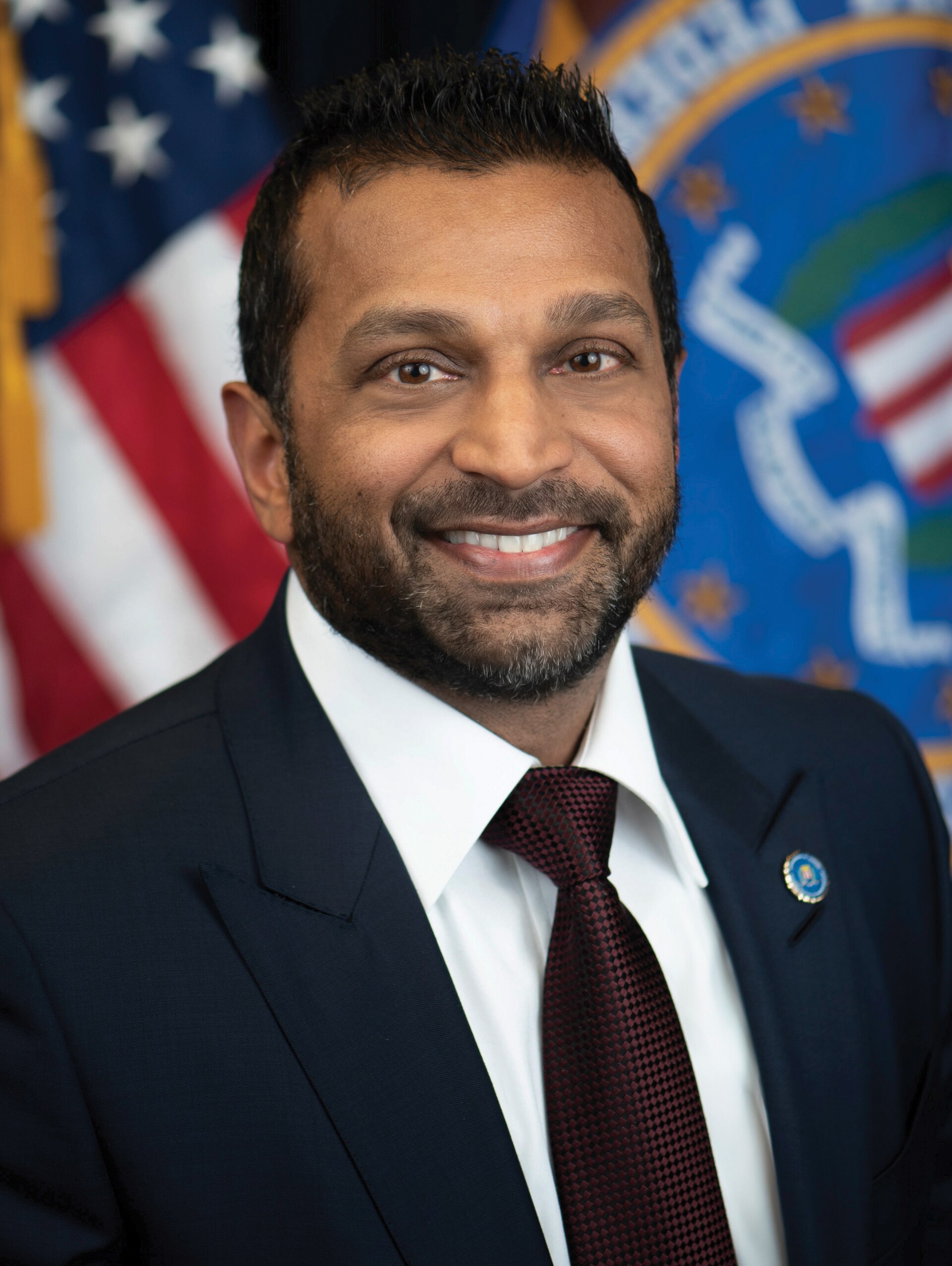 FBI Director Kash Patel smiles in a suit and tie, with the American flag and an FBI seal behind him.
