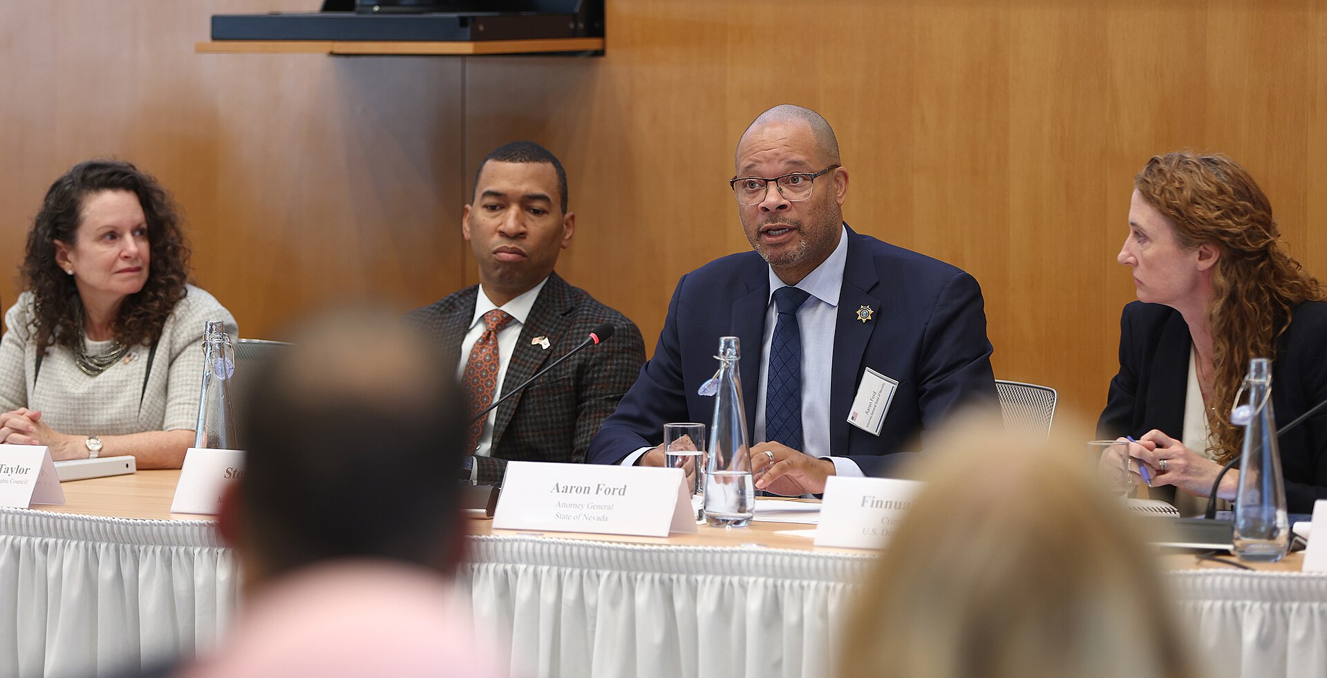 Aaron Ford, a bald man with glasses and a suit, sits at a table with a name card in front of him.