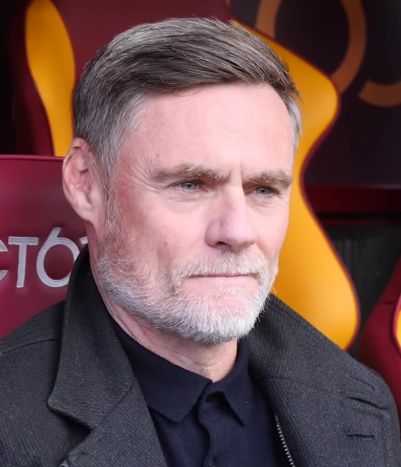 A man with short gray hair and a beard wearing a dark jacket and shirt