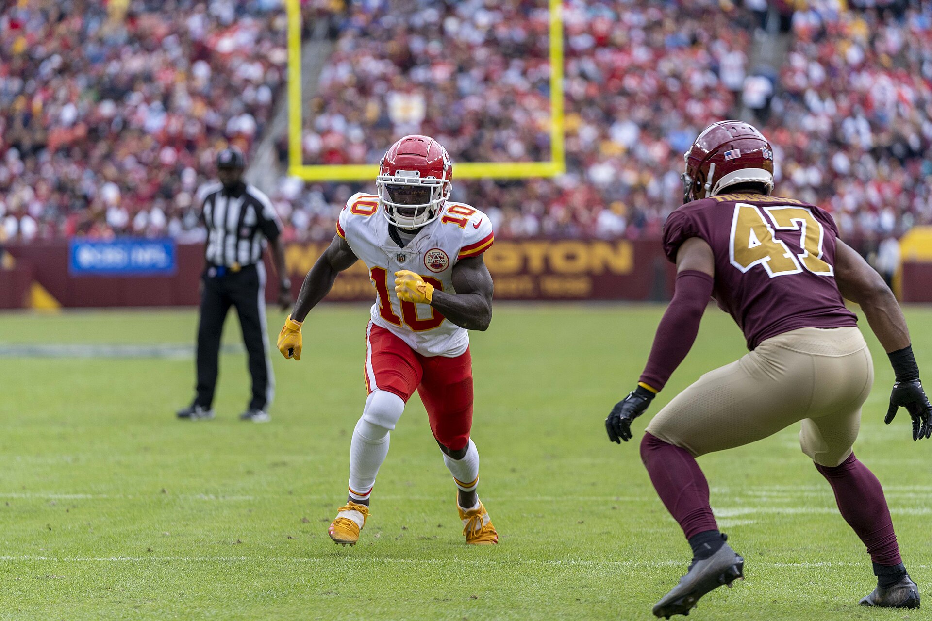 An American football player in a white and red uniform with the number 10 on the front runs on a green field, with another player in a maroon uniform with the number 47 on the back facing away from the camera. A referee in a black and white striped shirt stands in the background.