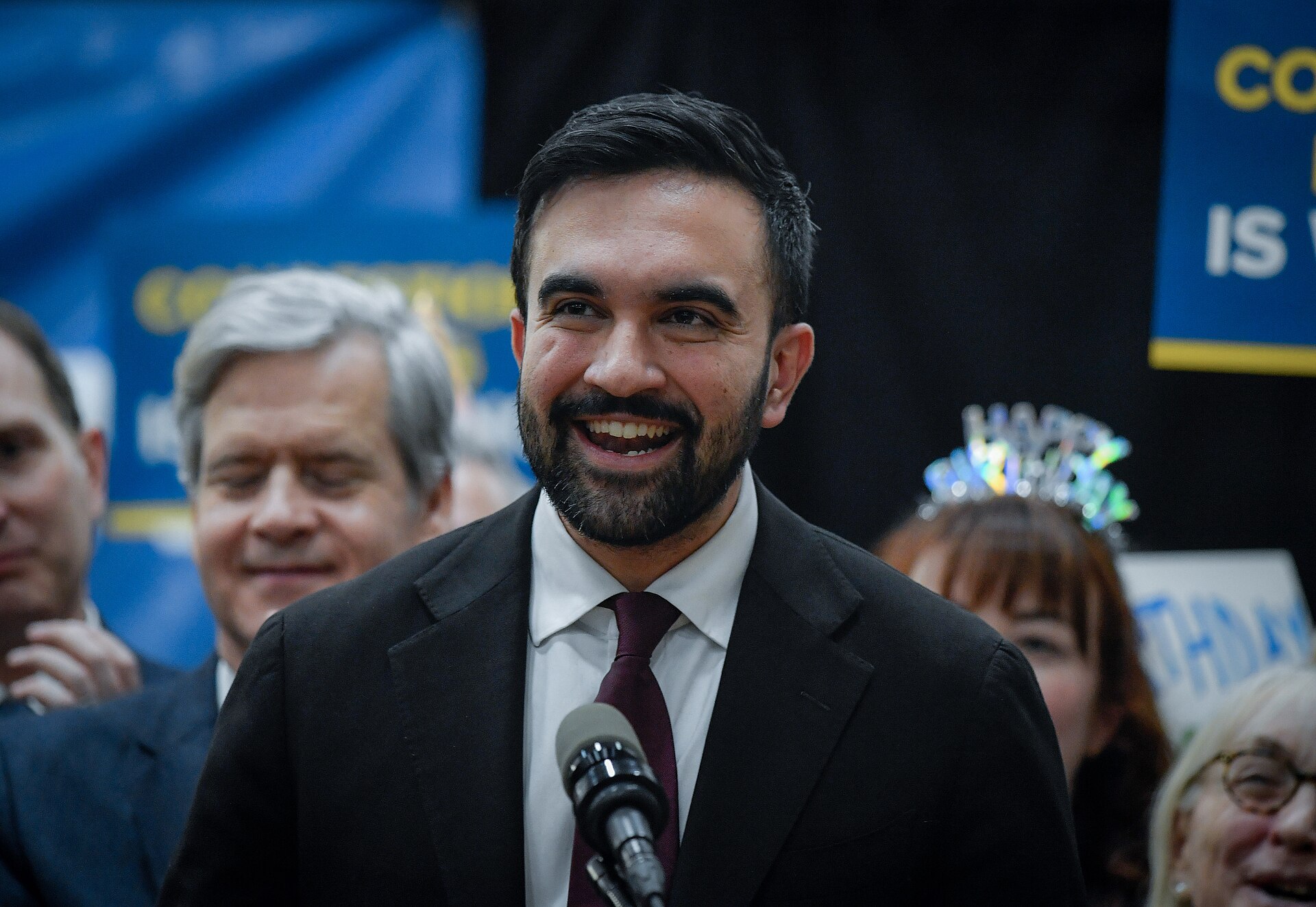 Zohran Mamdani, a bearded man in a black suit and maroon tie, speaks into a microphone, smiling, with a crowd behind him.
