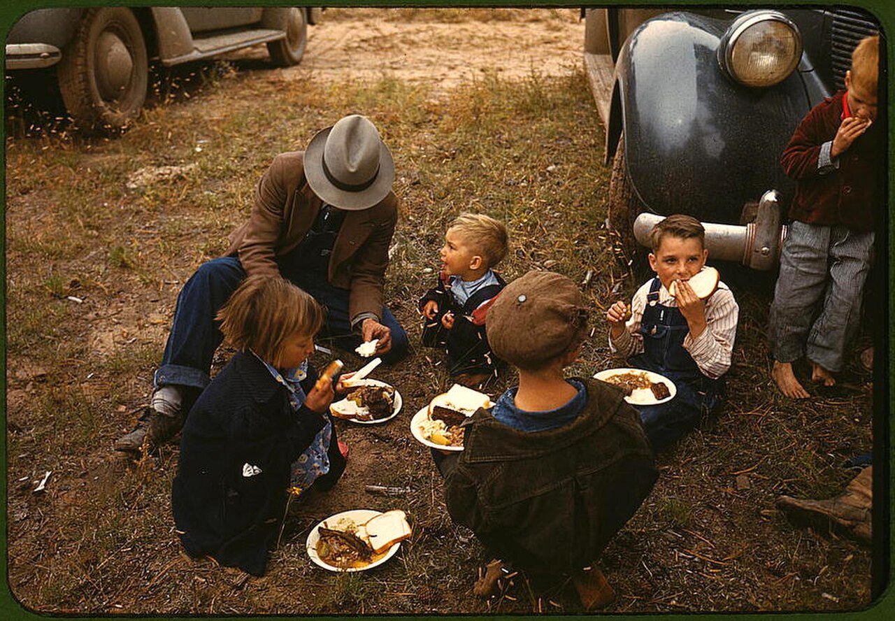 A group of people, including children and an adult man wearing a hat, are sitting on the ground eating meals from plates. They are gathered near a vintage car.