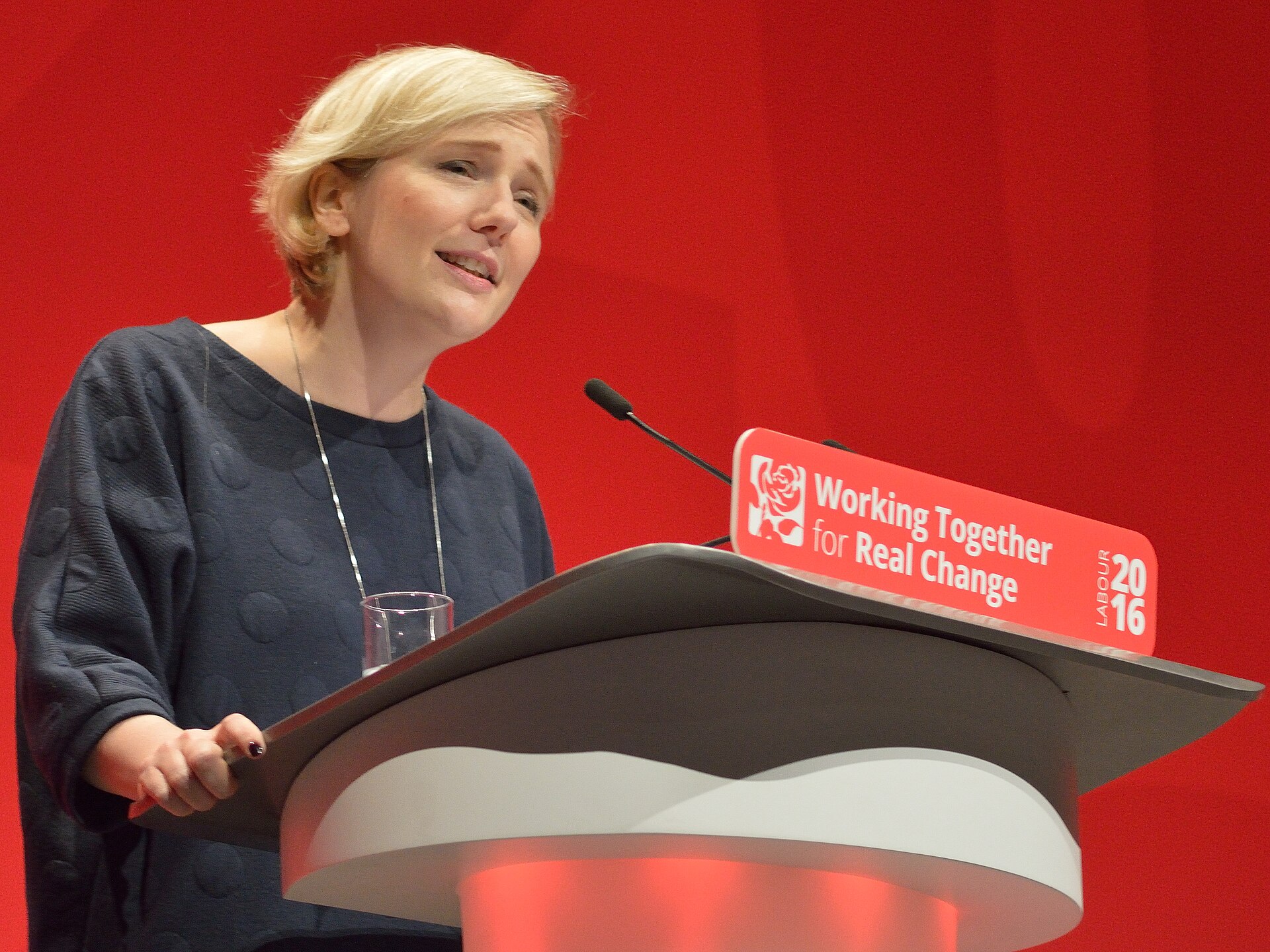A woman with short blonde hair, wearing a dark blue patterned sweater and a silver necklace, speaks at a podium. A red sign behind her reads "Working Together for Real Change" and "LABOUR 2016".