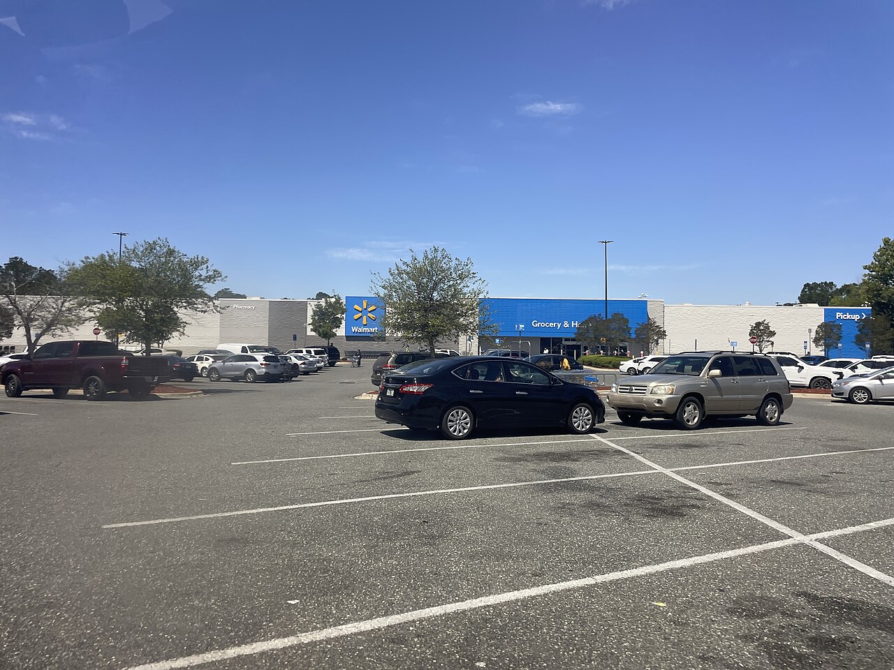 A Walmart Supercenter with a blue facade and a large blue Walmart logo is visible in the background, with a parking lot in the foreground. Cars are parked in the lot, and a few trees are scattered around the building.