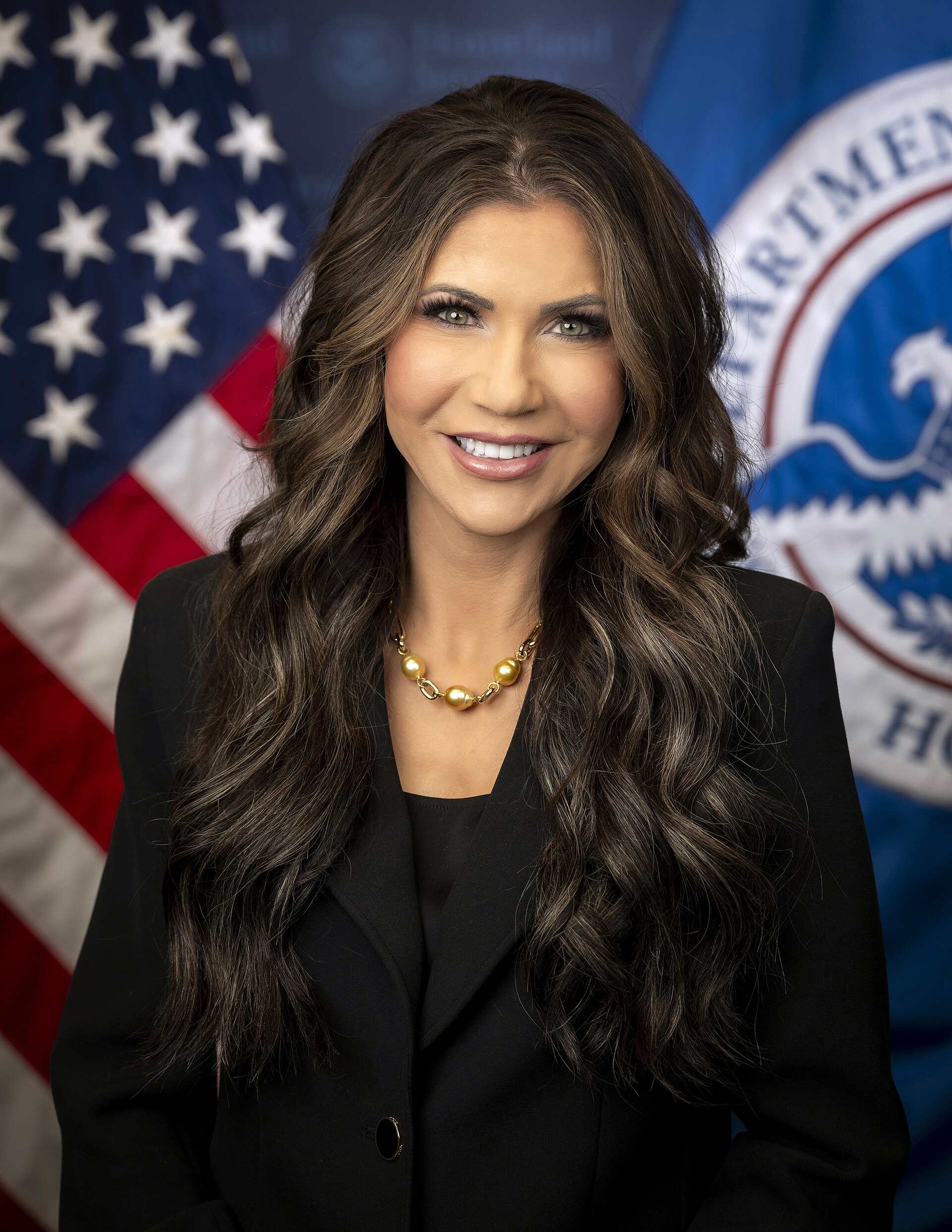 A smiling woman with long brown hair wearing a black suit and gold necklace, standing in front of an American flag and a blue flag with a seal.
