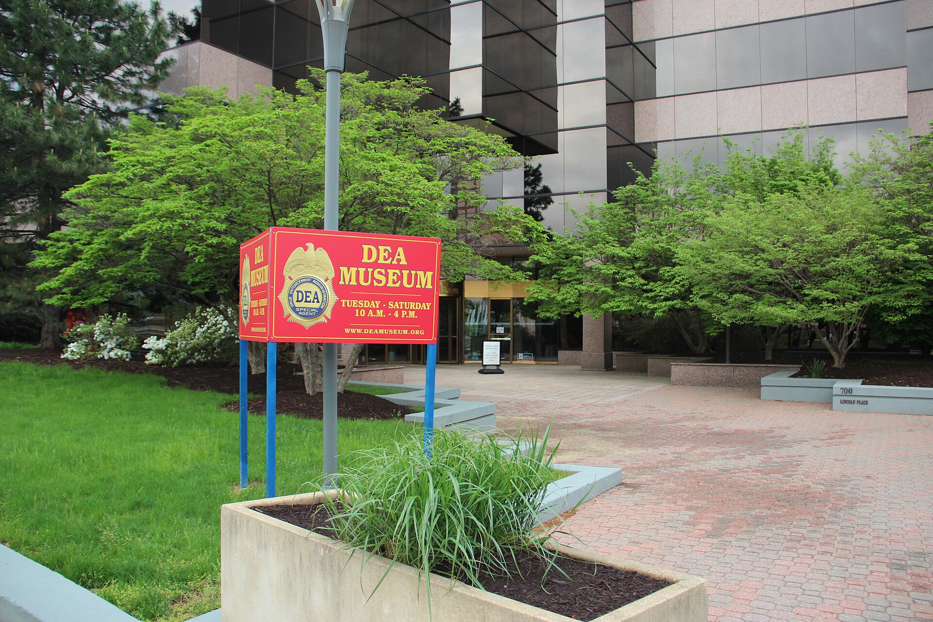 A red sign for the DEA Museum is visible in front of a modern building with reflective windows. The sign indicates the museum is open Tuesday through Saturday from 10 AM to 4 PM and provides a website address.