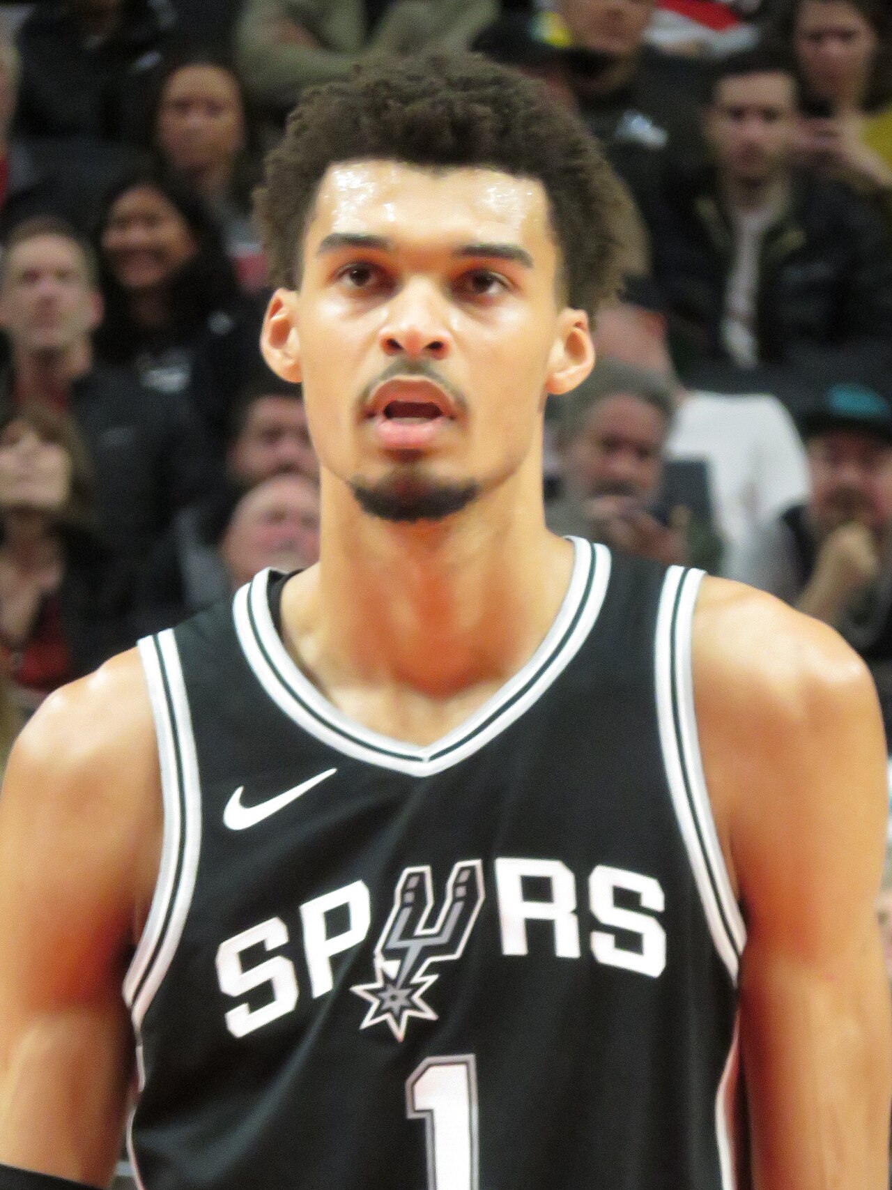 A young man with short dark hair wearing a black basketball jersey with white trim and the word SPURS on the front
