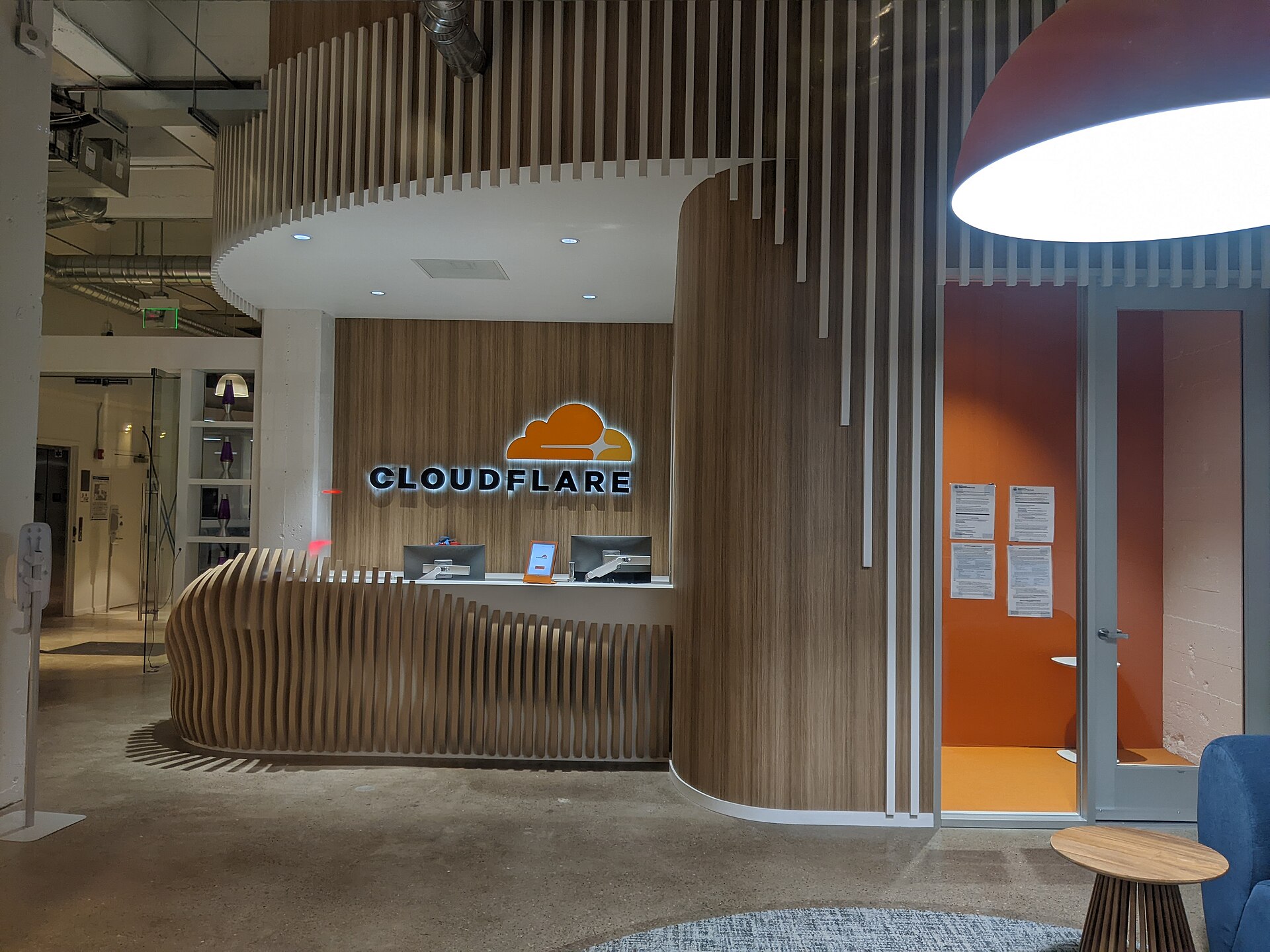 The Cloudflare logo is displayed above a reception desk in a modern office lobby. The logo features an orange cloud with a white outline, next to the word "CLOUDFLARE" in white capital letters. The reception desk has a curved wooden front and a white countertop with two computer monitors and a table