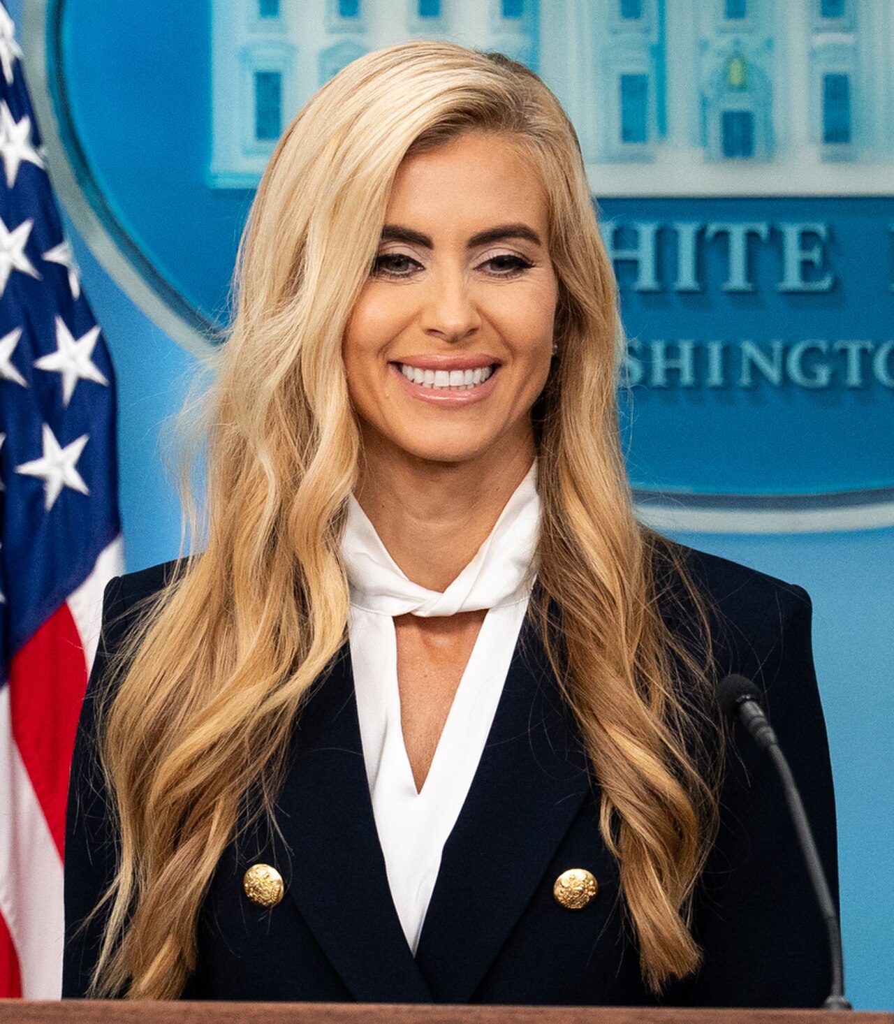 A blonde woman in a dark blue blazer and white blouse smiles at a podium in front of a White House backdrop and an American flag.
