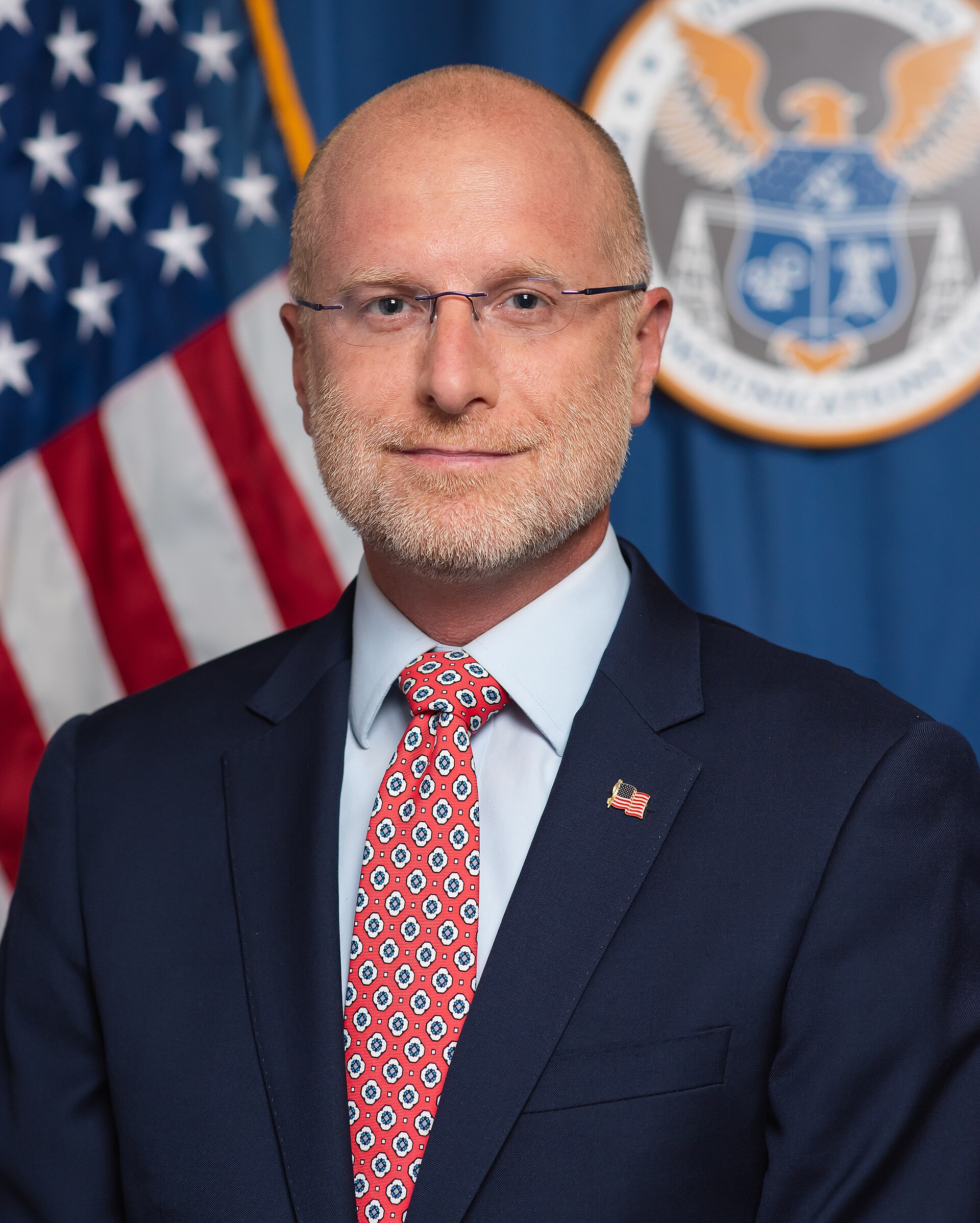 A man in a suit and tie stands in front of an American flag and FCC logo, wearing a small American flag pin on his lapel.