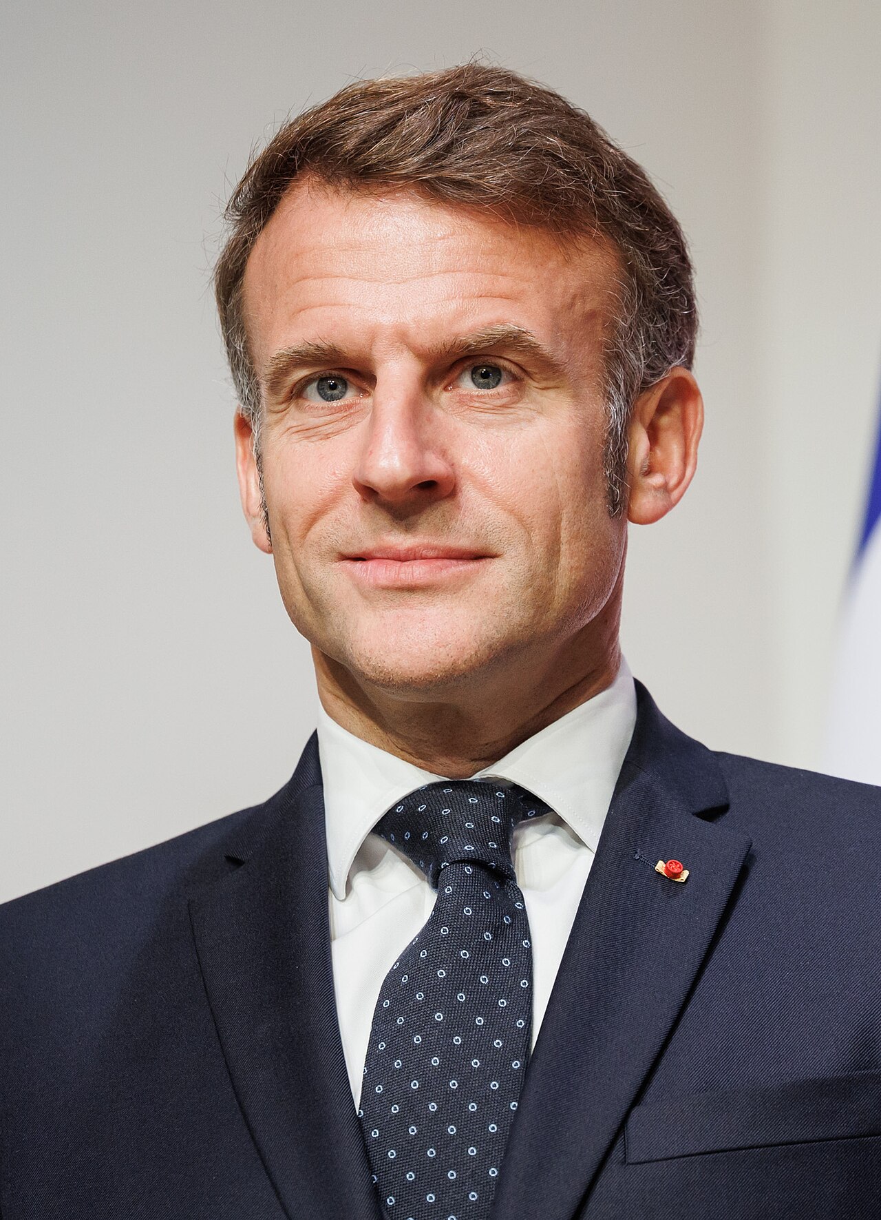 Emmanuel Macron, wearing a dark blue suit, white shirt, and dark blue tie with small white dots, looking directly at the camera.