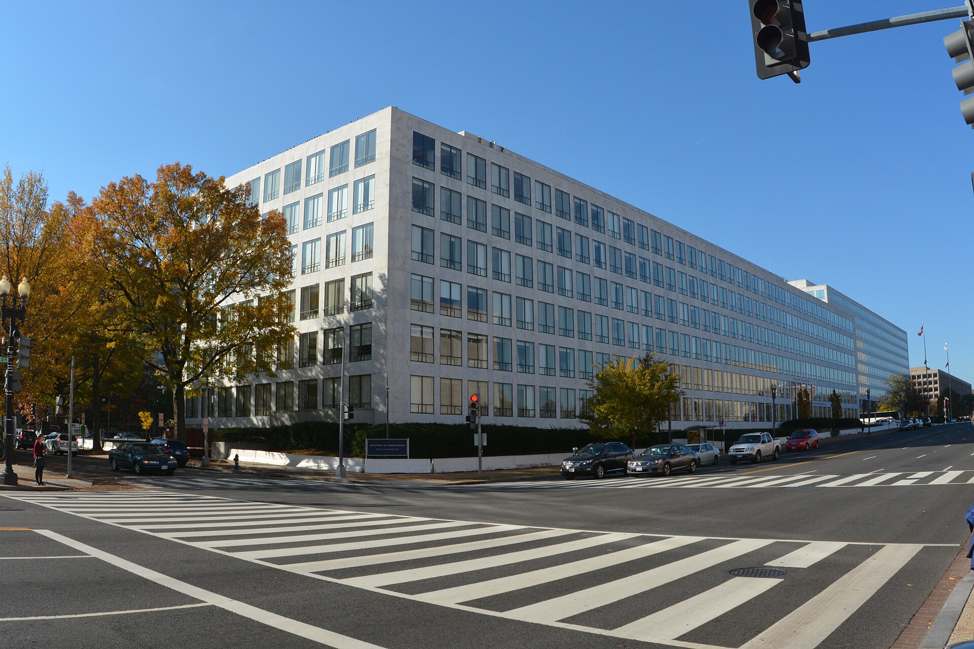 A wide, modern, light-colored building with many windows, identified by a sign as the Department of Transportation, stands on a street corner with a crosswalk in the foreground and trees with autumn leaves.