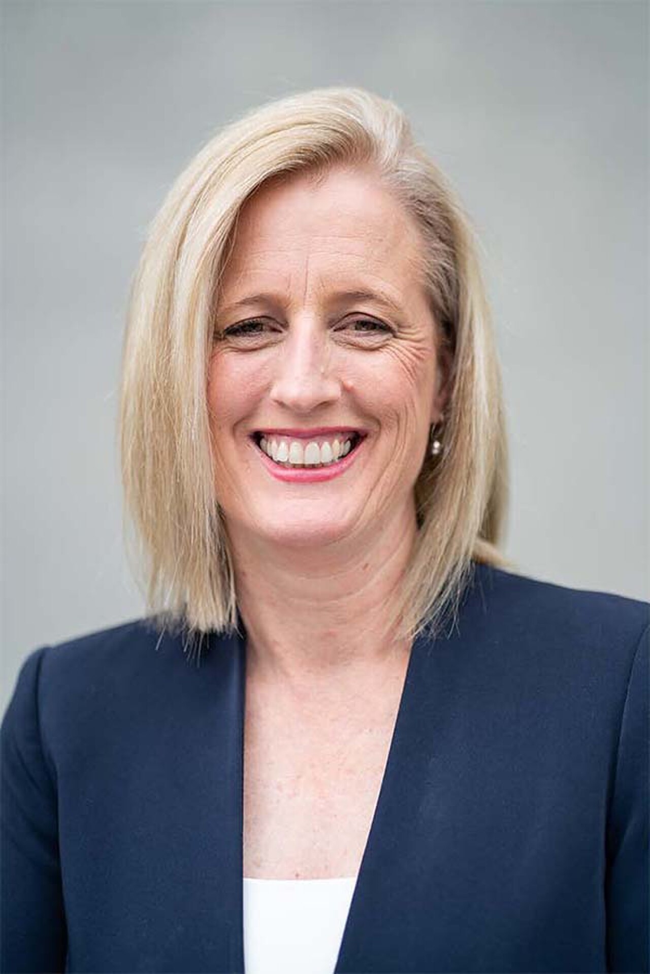 A smiling middle-aged woman with shoulder-length blonde hair wearing a navy blue blazer and a white top, against a light gray background.