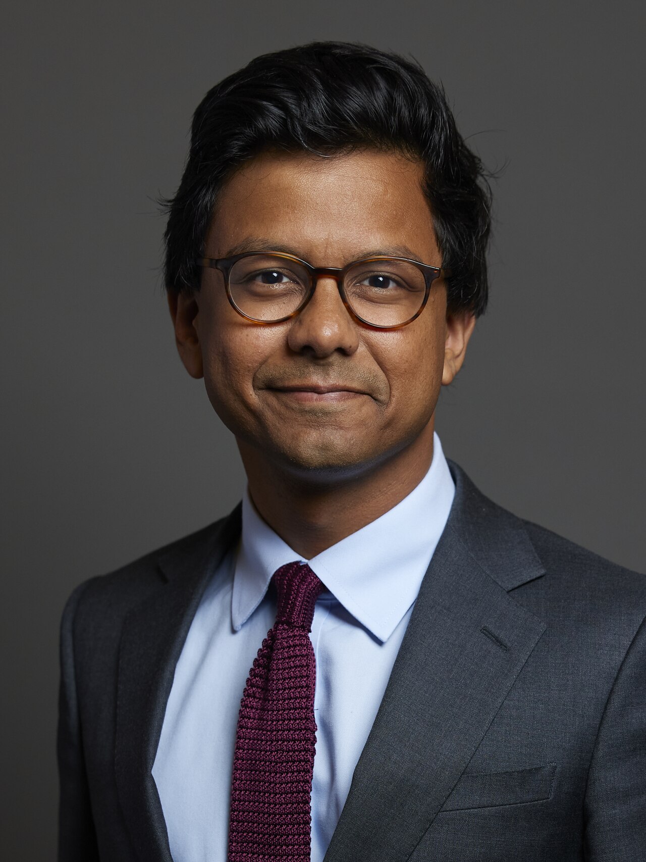 A brown skinned man with black hair and glasses wearing a suit and tie