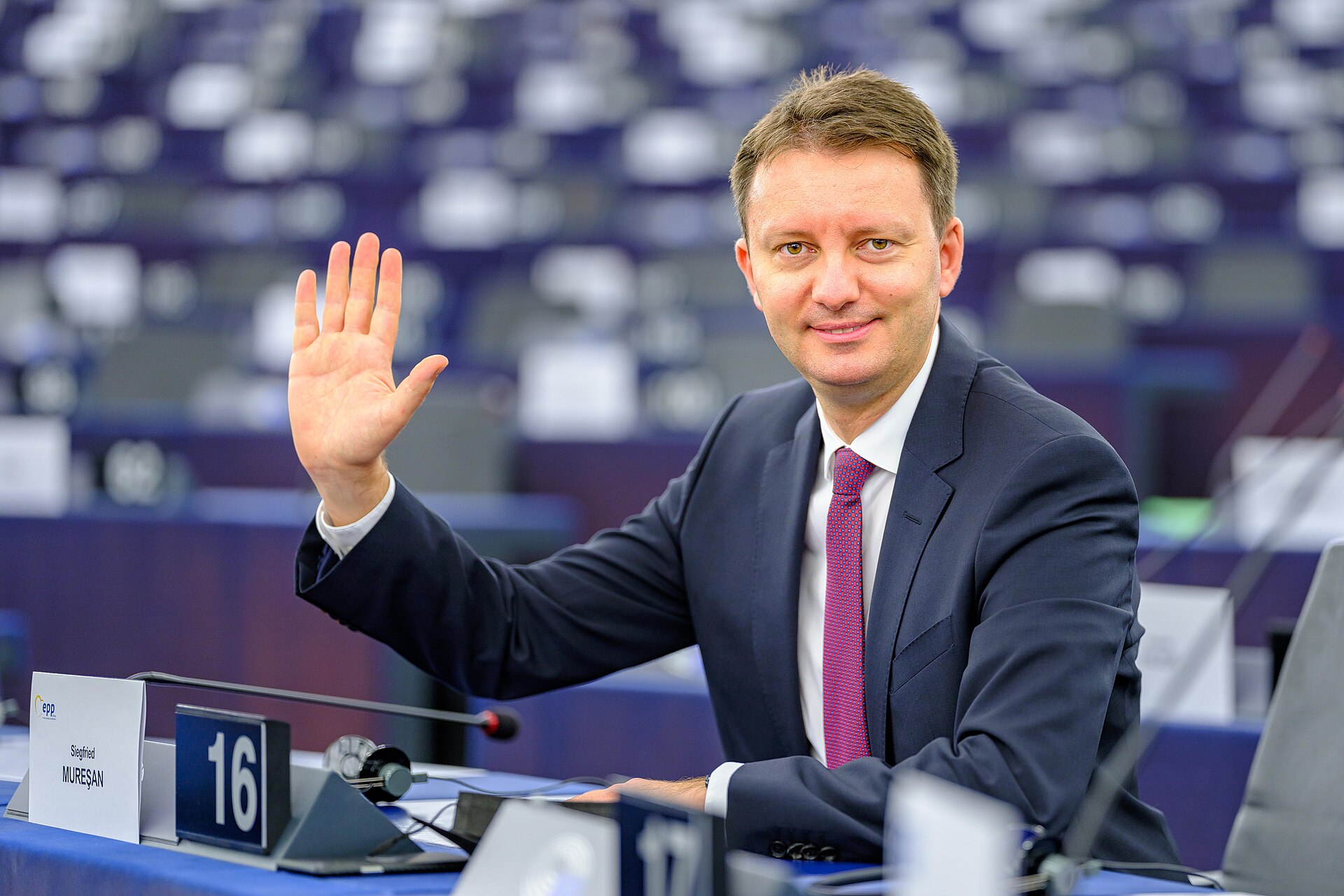 A man in a suit with his right hand raised, sitting at a desk with a name card reading 'Siegfried Muresan' and a number 16.
