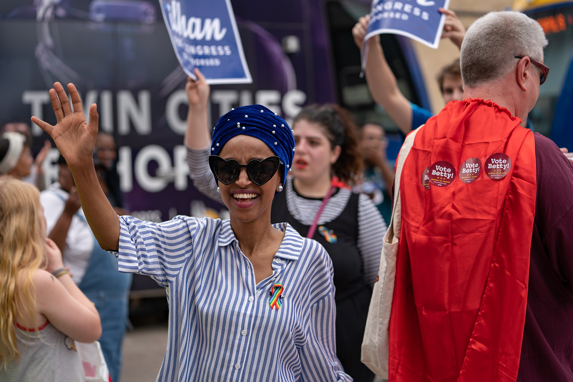 Ilhan Omar, wearing a blue headscarf, heart-shaped sunglasses, and a blue and white striped shirt with a rainbow ribbon, waves to a crowd. Behind her, people hold signs that say "Ilhan for Congress."