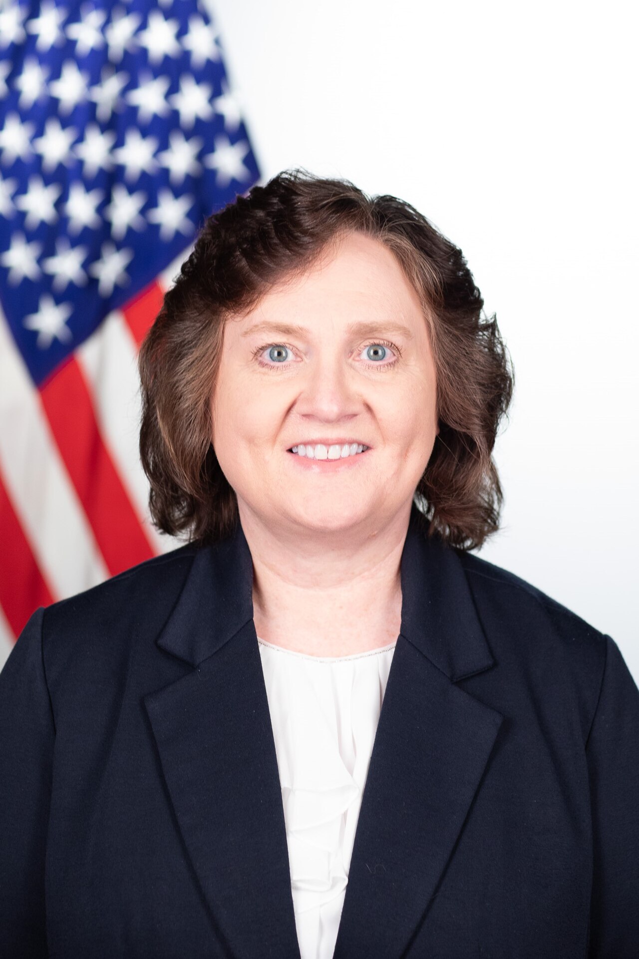 A woman with short brown hair, blue eyes, and a black blazer, smiling in front of a blurred American flag.