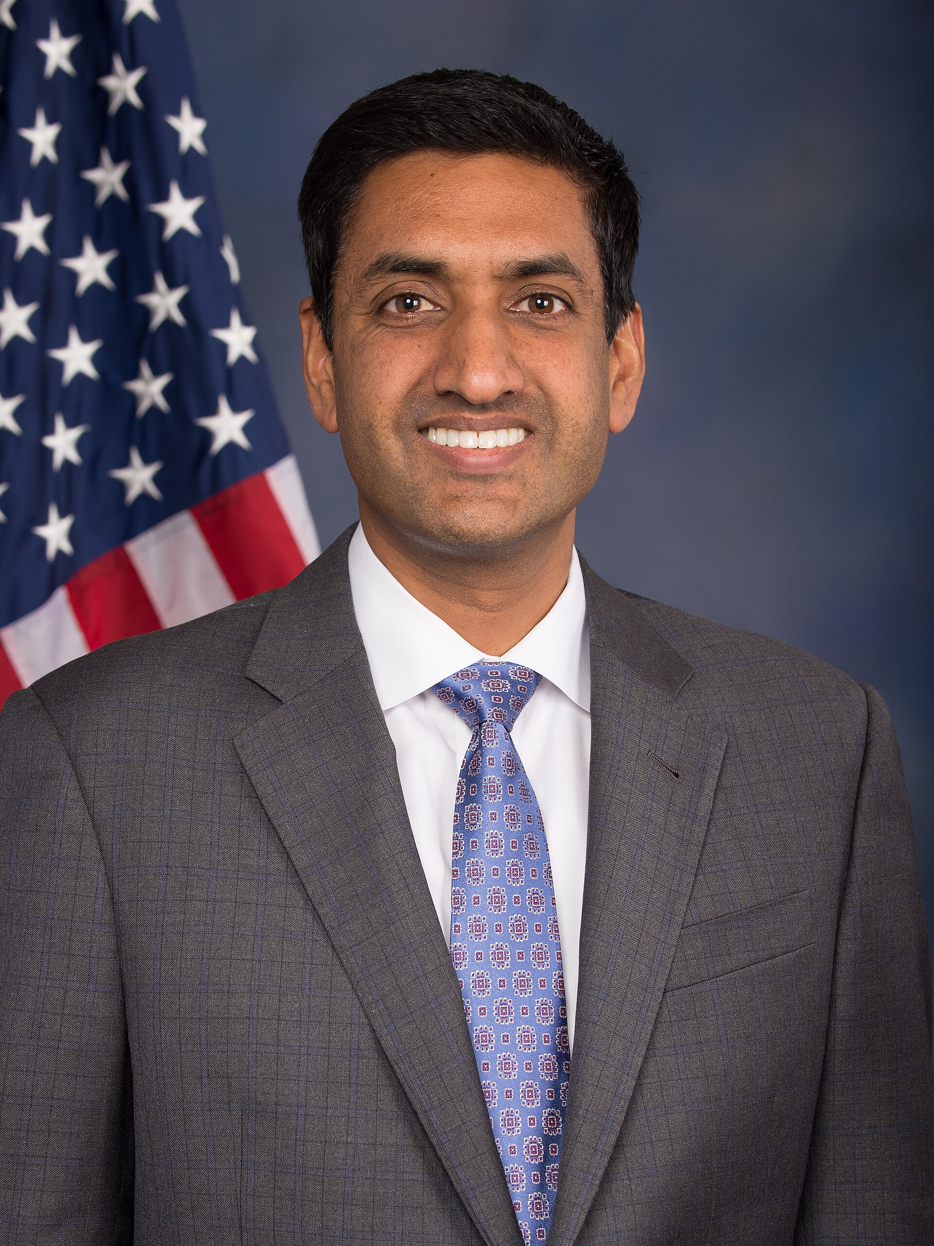 A man with light brown skin and short black hair, wearing a gray suit, white shirt, and blue patterned tie, stands in front of a blue background with an American flag on the left.