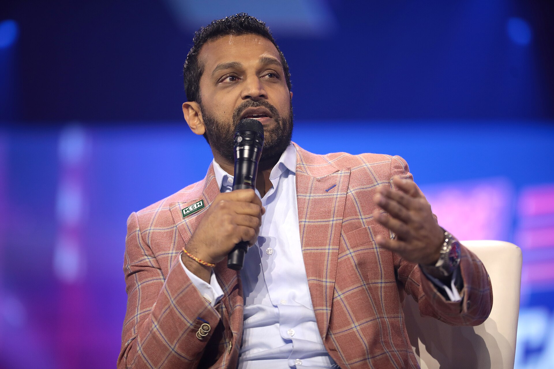 A man with light brown skin and short dark hair, wearing a pink plaid suit jacket, white shirt, and holding a microphone, gesturing with his left hand while seated.