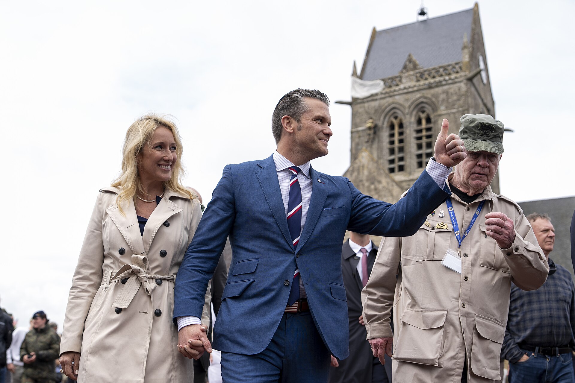 Pete Hegseth in a blue suit pointing outward, holding a woman's hand, walking with a man in a tan uniform and cap