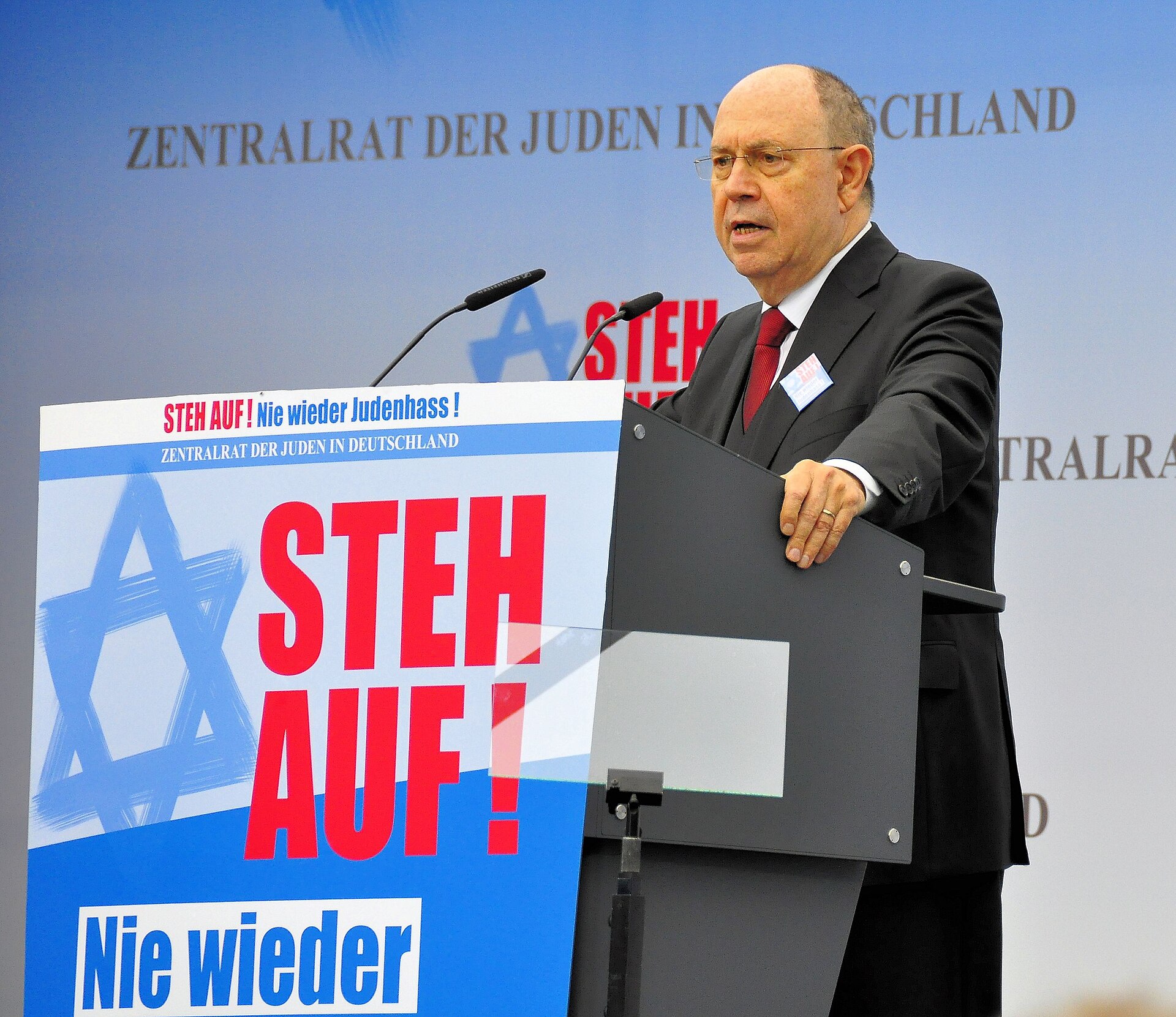 A man in a suit and tie speaks at a podium with a banner behind him that reads "STEH AUF! Nie wieder Judenhass!" and "ZENTRALRAT DER JUDEN IN DEUTSCHLAND".
