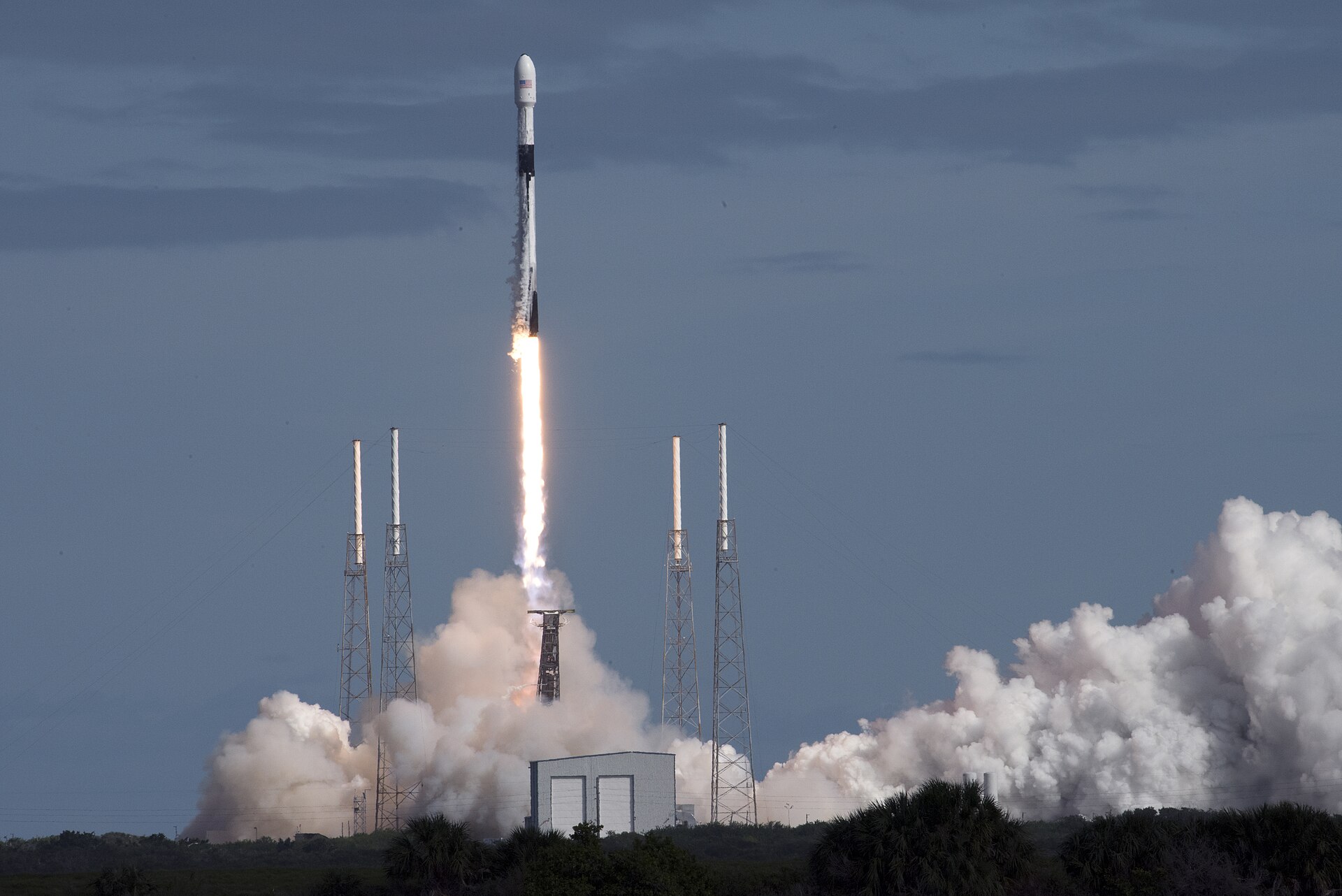 A rocket launches into the sky, trailing smoke and flames, with launch towers and a building visible on the ground.